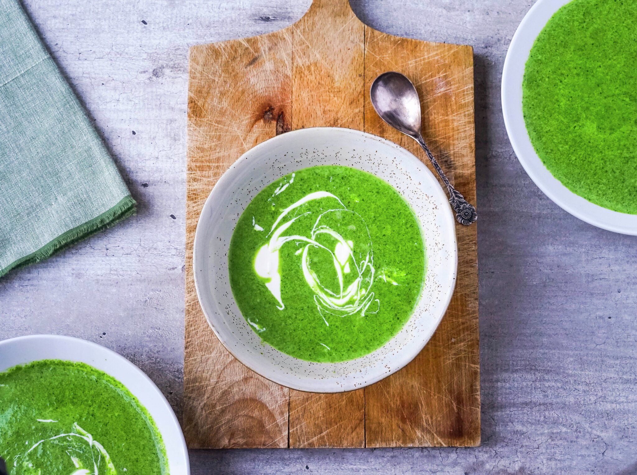 Three bowls of vibrant green soup, two garnished with white cream swirls, served on a wooden board and gray table with a spoon.