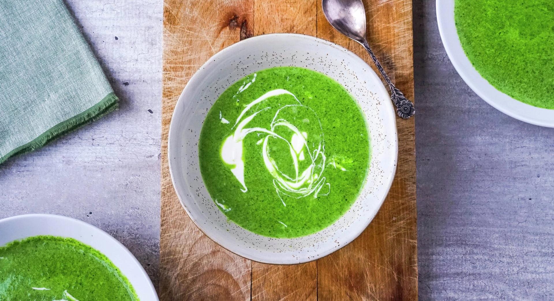 Three bowls of vibrant green soup, two garnished with white cream swirls, served on a wooden board and gray table with a spoon.