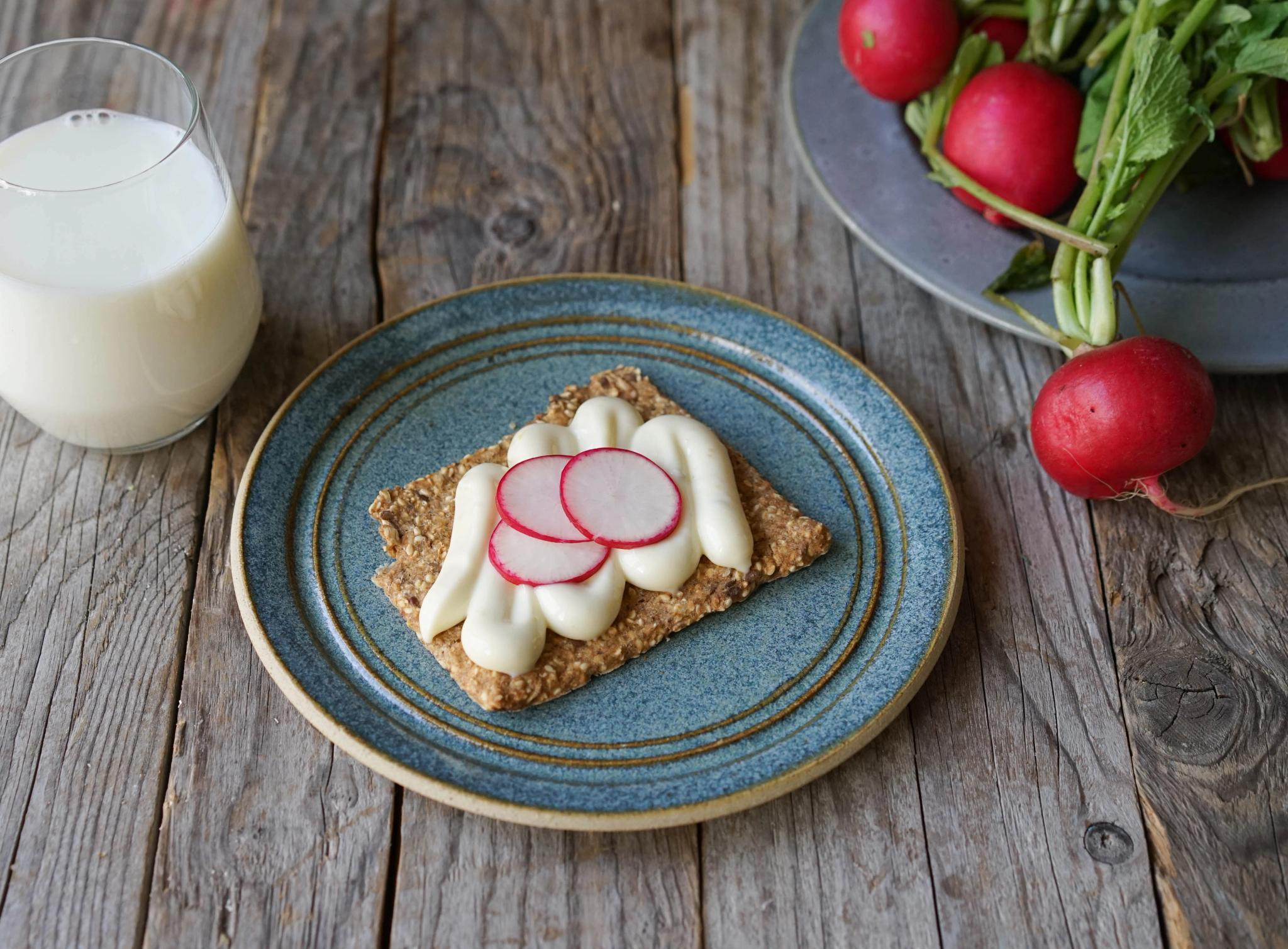 Crispbread with cream and radish slices on a blue plate, next to a glass of milk and fresh radishes on a wooden table.