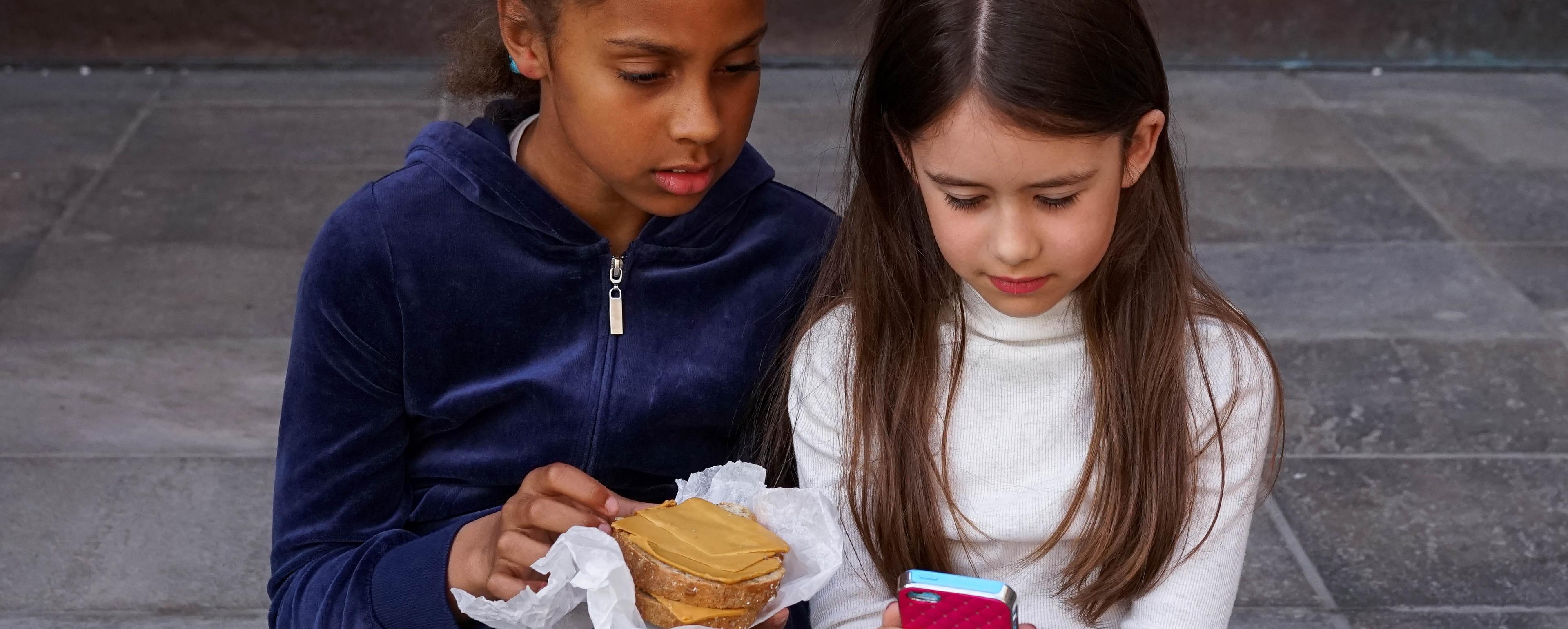To jenter ser på mobil og spiser matpakke