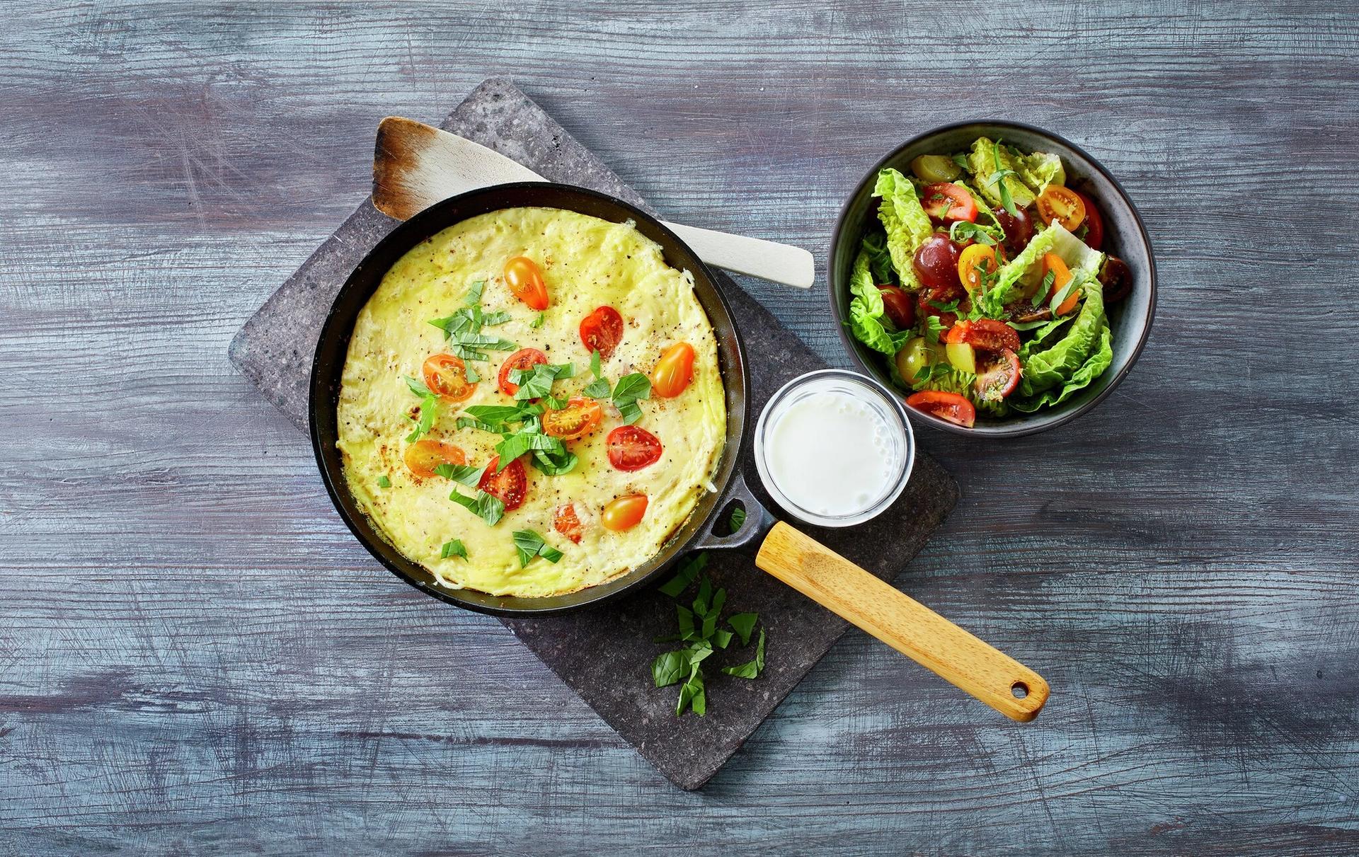 Omelett med tomater i en stekepanne med et glass melk og en skål salat ved siden