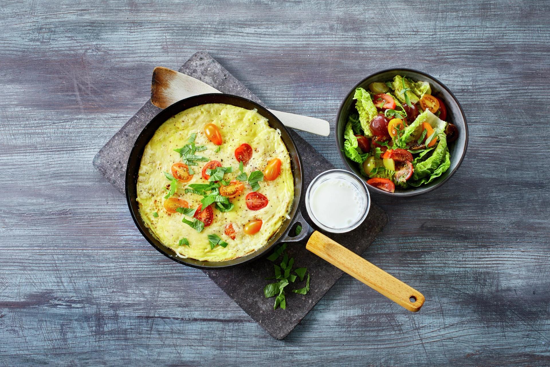 Omelett med tomater i en stekepanne med et glass melk og en skål salat ved siden