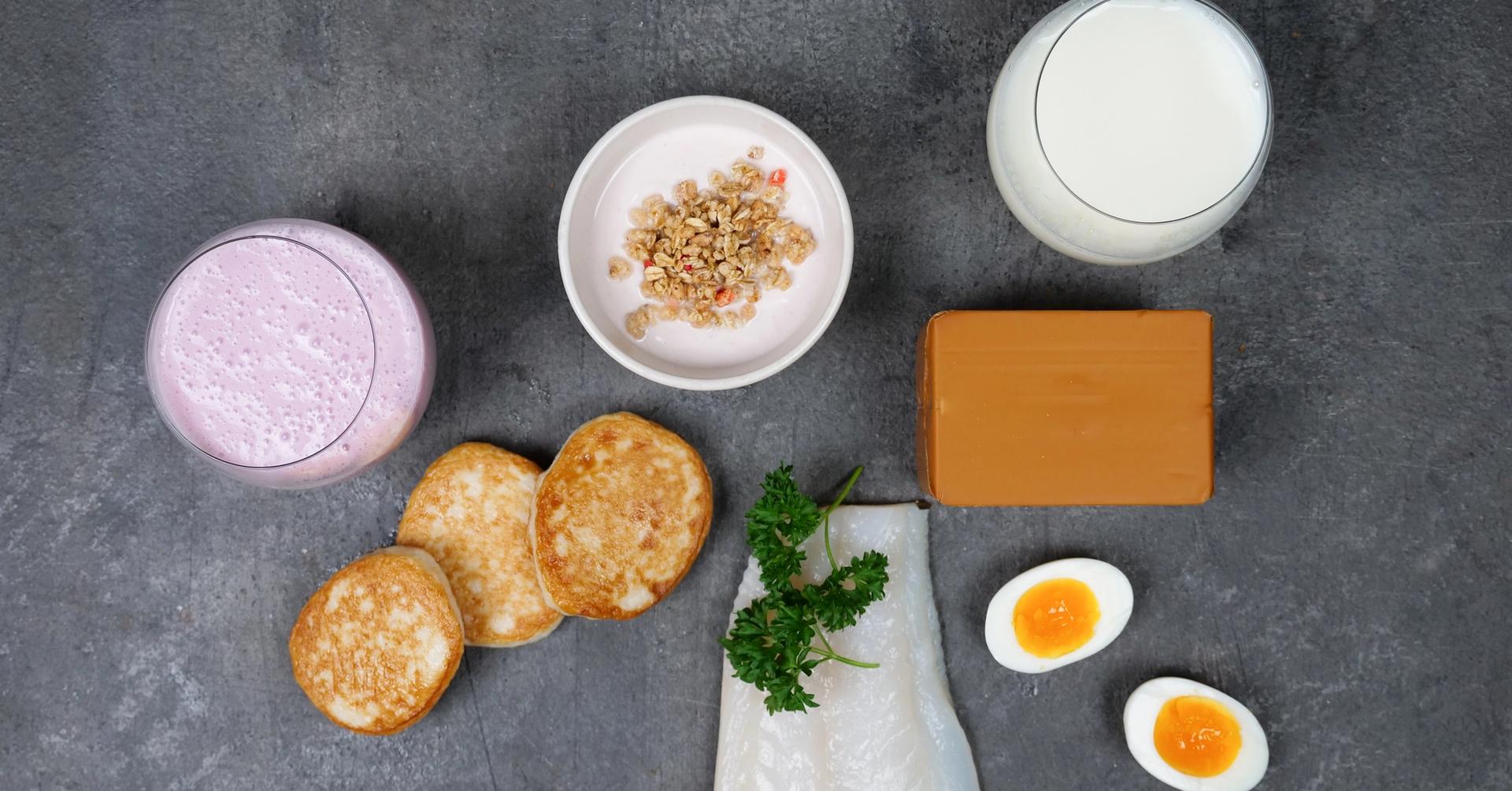 A variety of food items including a pink smoothie, granola, pancakes, brown cheese, milk, white fish with parsley, and two halved boiled eggs on a dark surface.