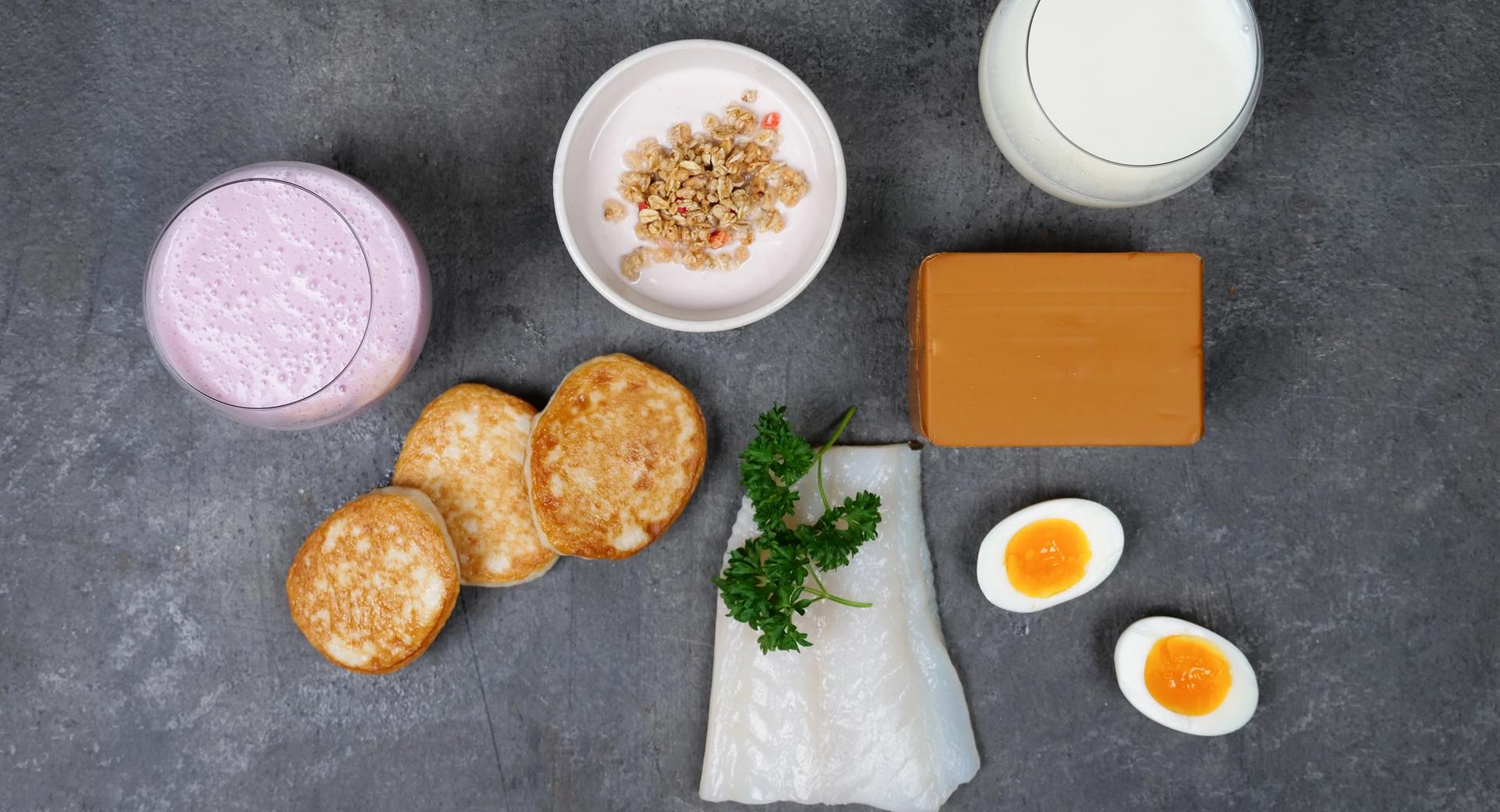 A variety of food items including a pink smoothie, granola, pancakes, brown cheese, milk, white fish with parsley, and two halved boiled eggs on a dark surface.