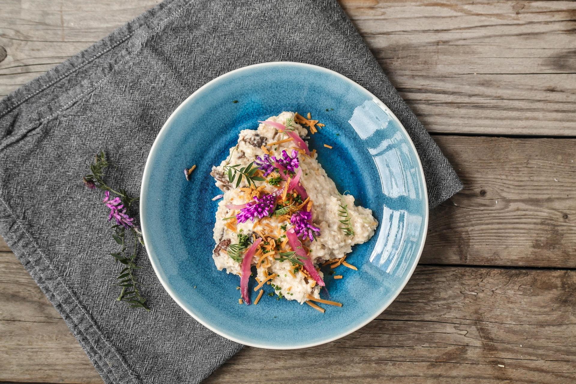 A creamy dish garnished with purple flowers, pickled red onions, and fried shallots on a blue plate, on a wooden table.