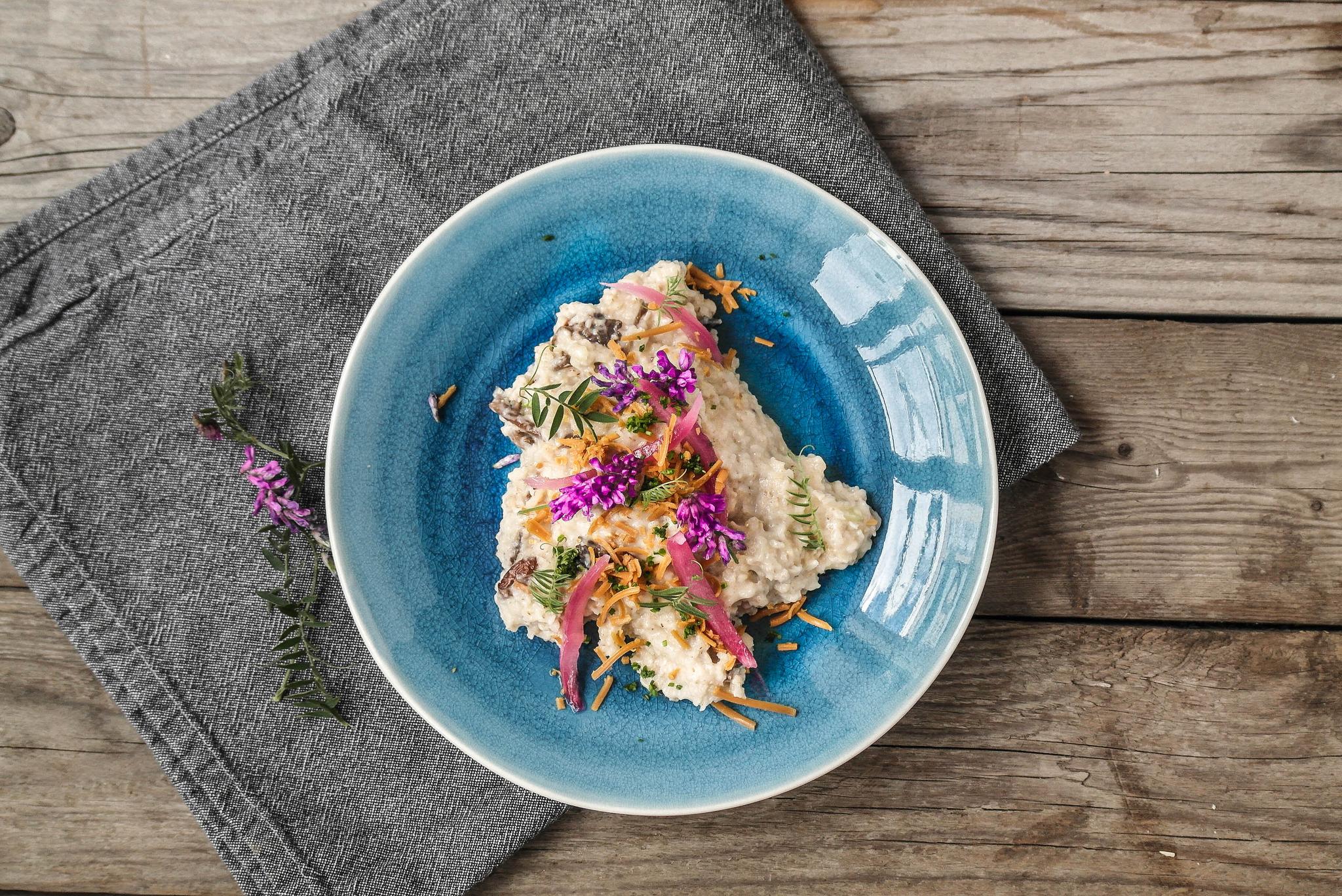 A creamy dish garnished with purple flowers, pickled red onions, and fried shallots on a blue plate, on a wooden table.