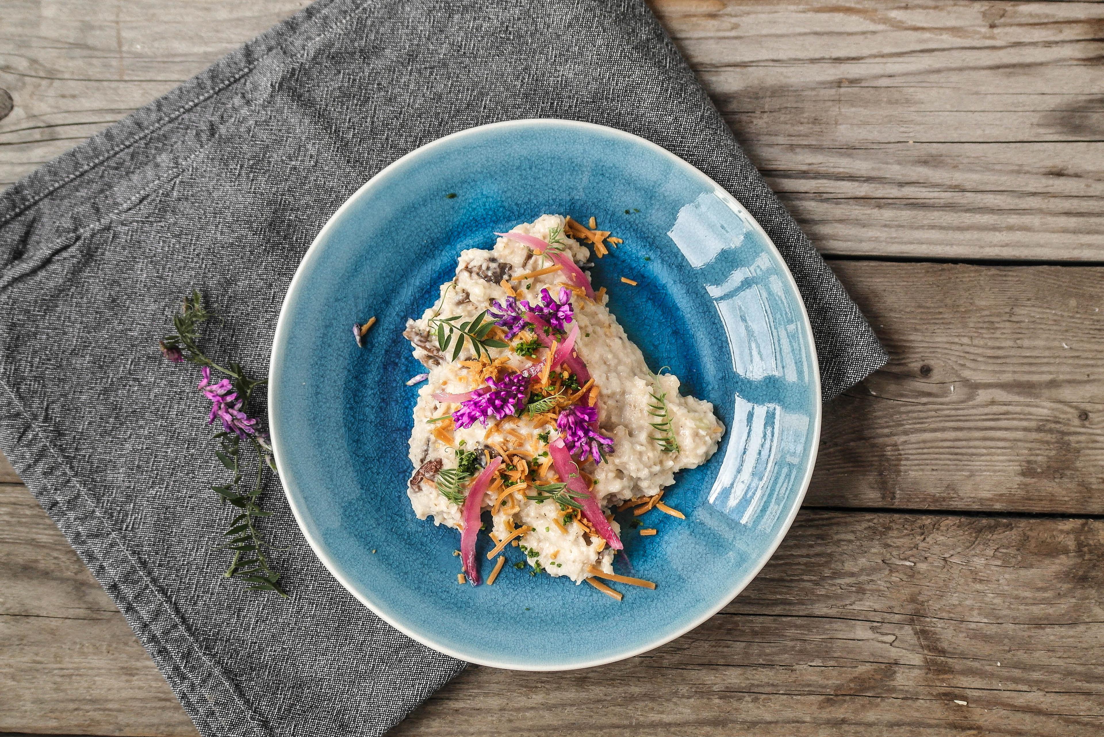 A creamy dish garnished with purple flowers, pickled red onions, and fried shallots on a blue plate, on a wooden table.