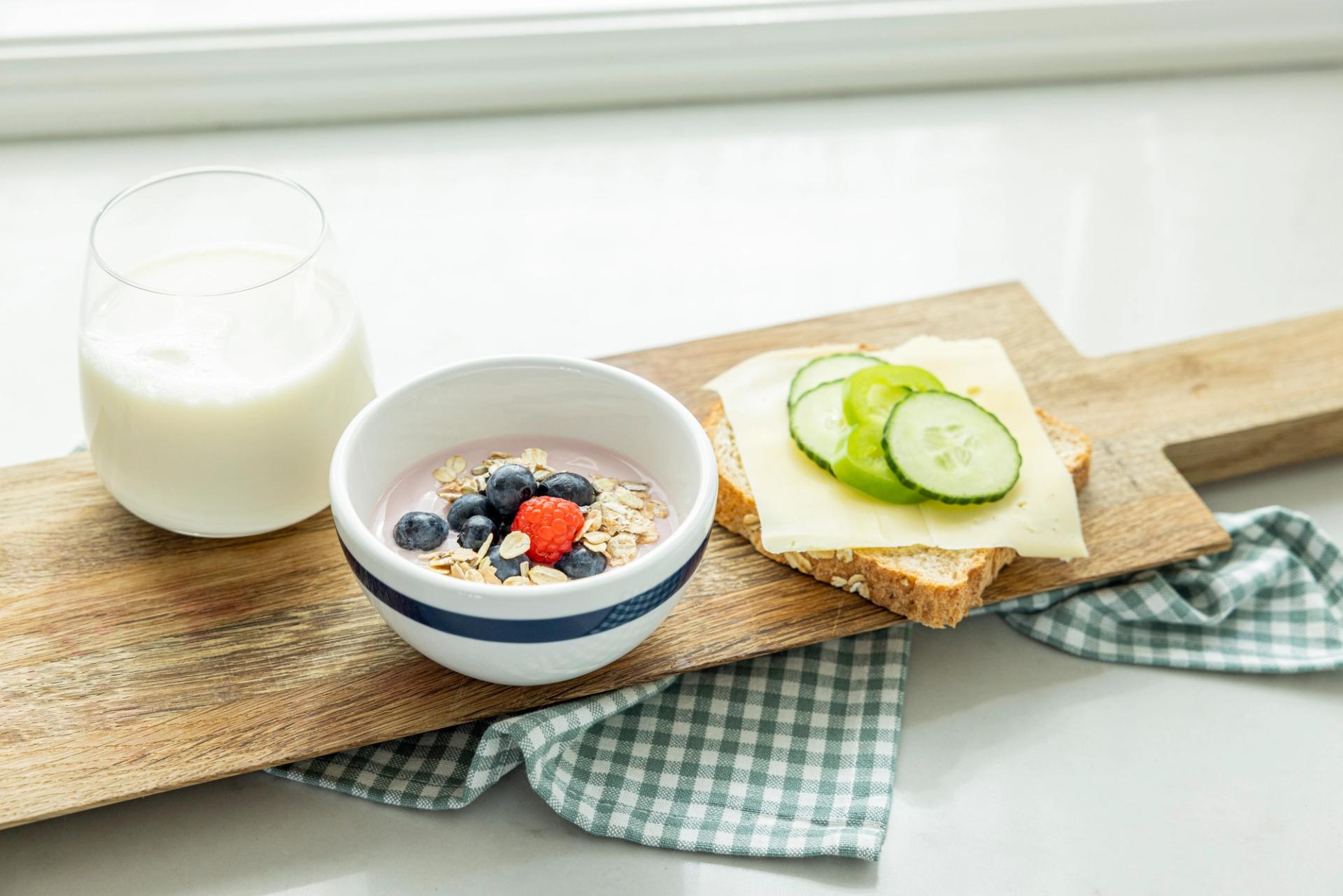 Fjøl med et glass melk, yoghurt med korn og bær og en brødskive med gulost og agurkskiver