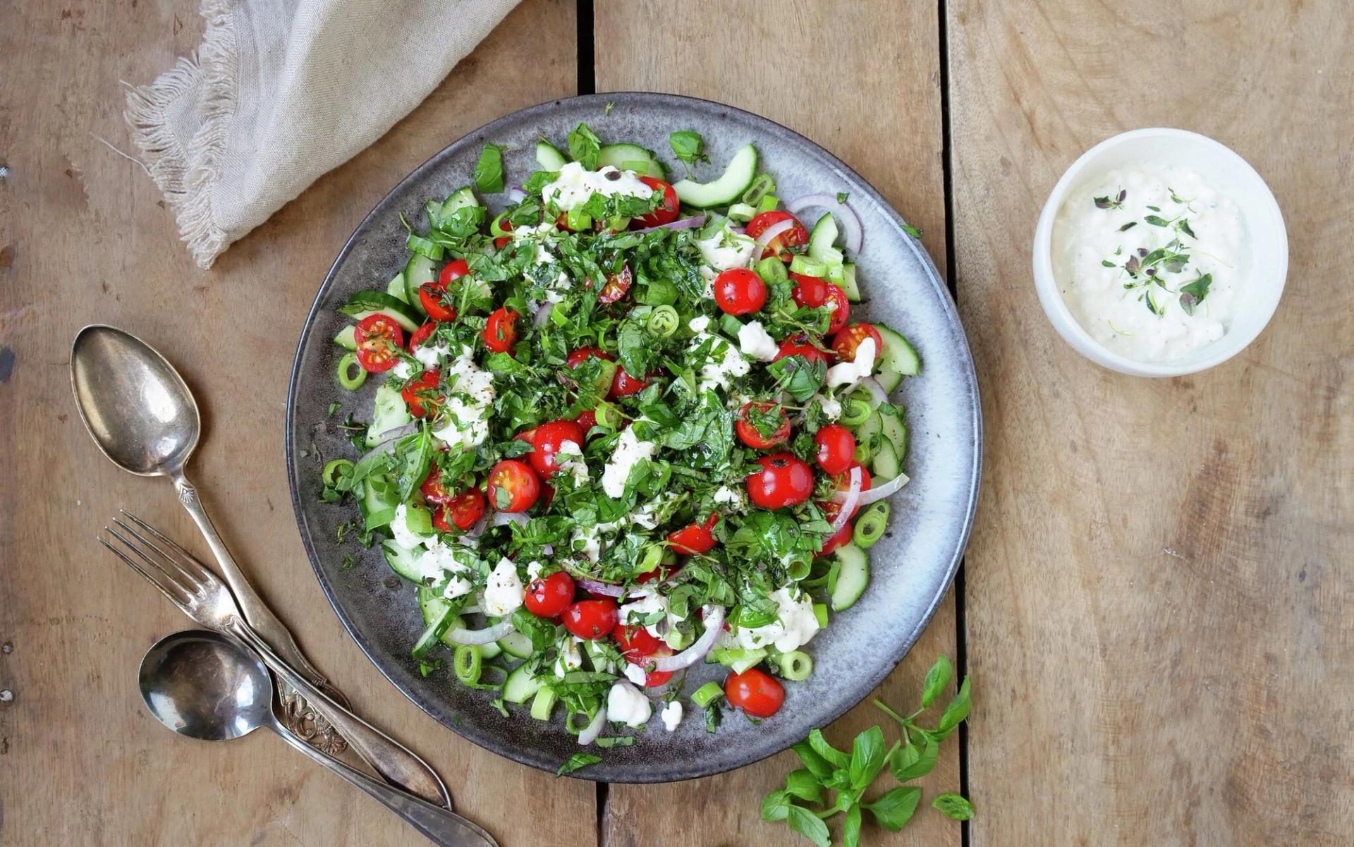 Vibrant salad with cherry tomatoes, cucumber, feta, and fresh herbs on a rustic wooden table with silverware and a dip.