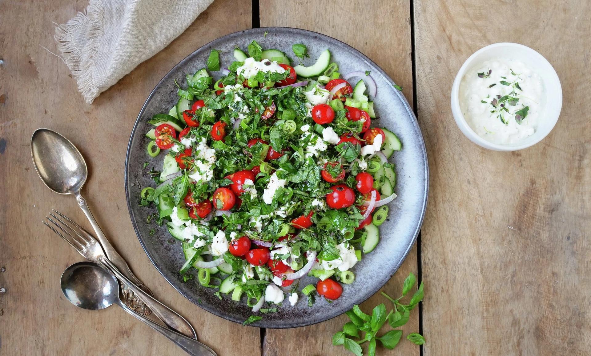 Bolle med salat med tomater, agurk og cottage cheese. To skjeer, en gaffel og en skål med hvit dressing.