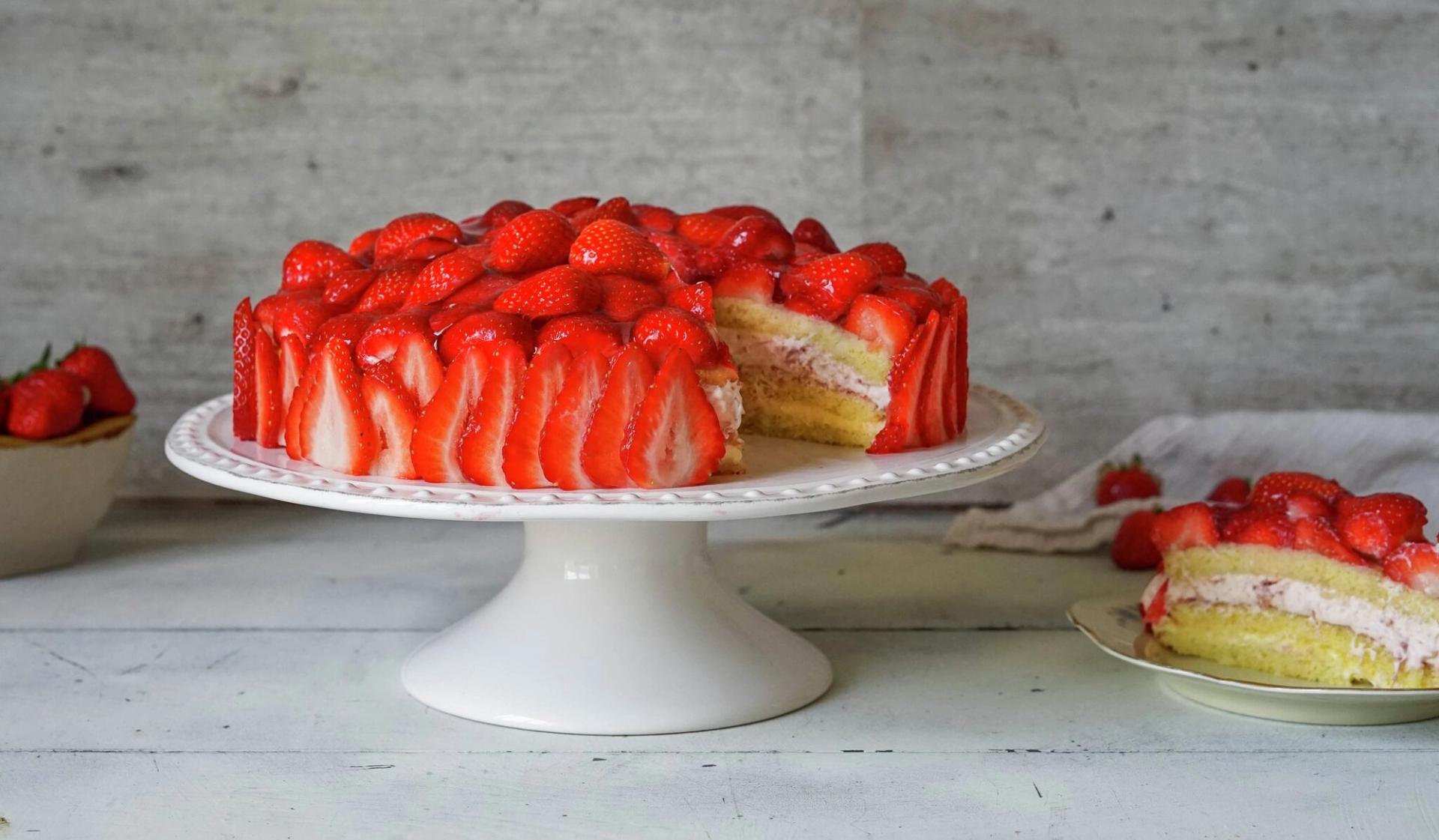 A strawberry cake on a white cake stand, decorated with sliced strawberries on the sides and whole strawberries on top. A cut slice reveals yellow cake and pink cream layers, resting on a small plate.