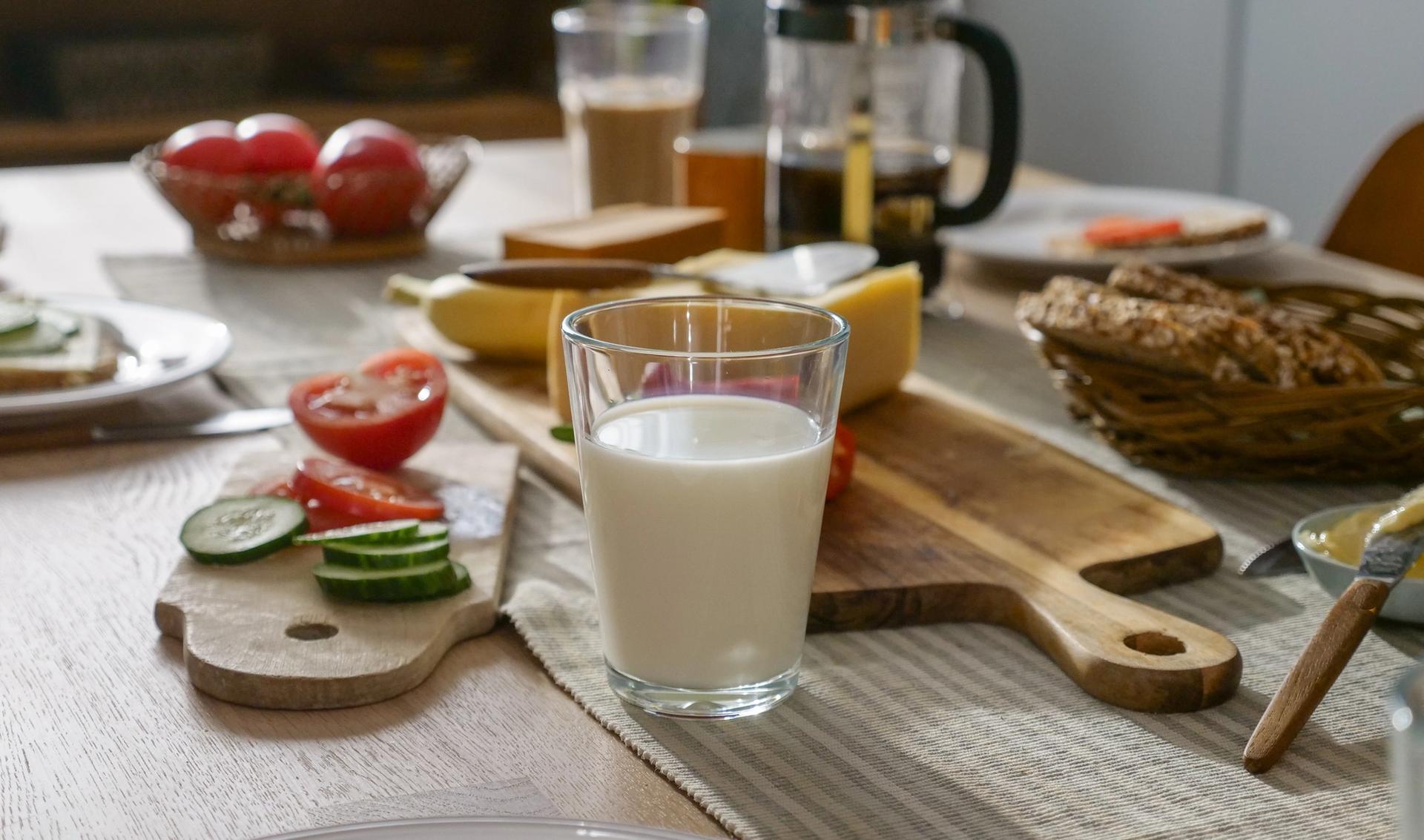 a glass of milk is sitting on a wooden table next to a cutting board with vegetables .