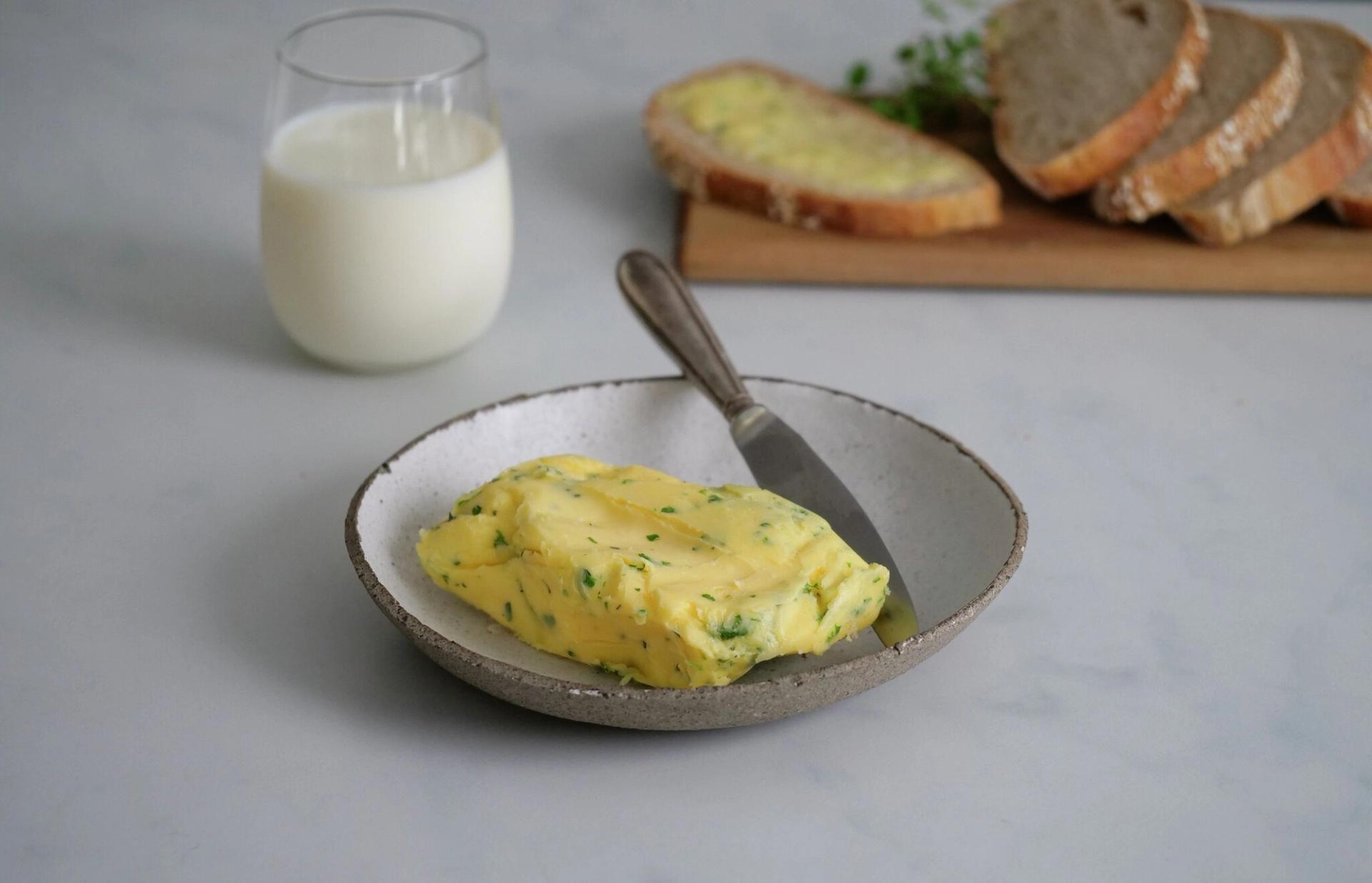 A block of herbed butter with a knife in a bowl, a glass of milk, and sliced bread in the background.