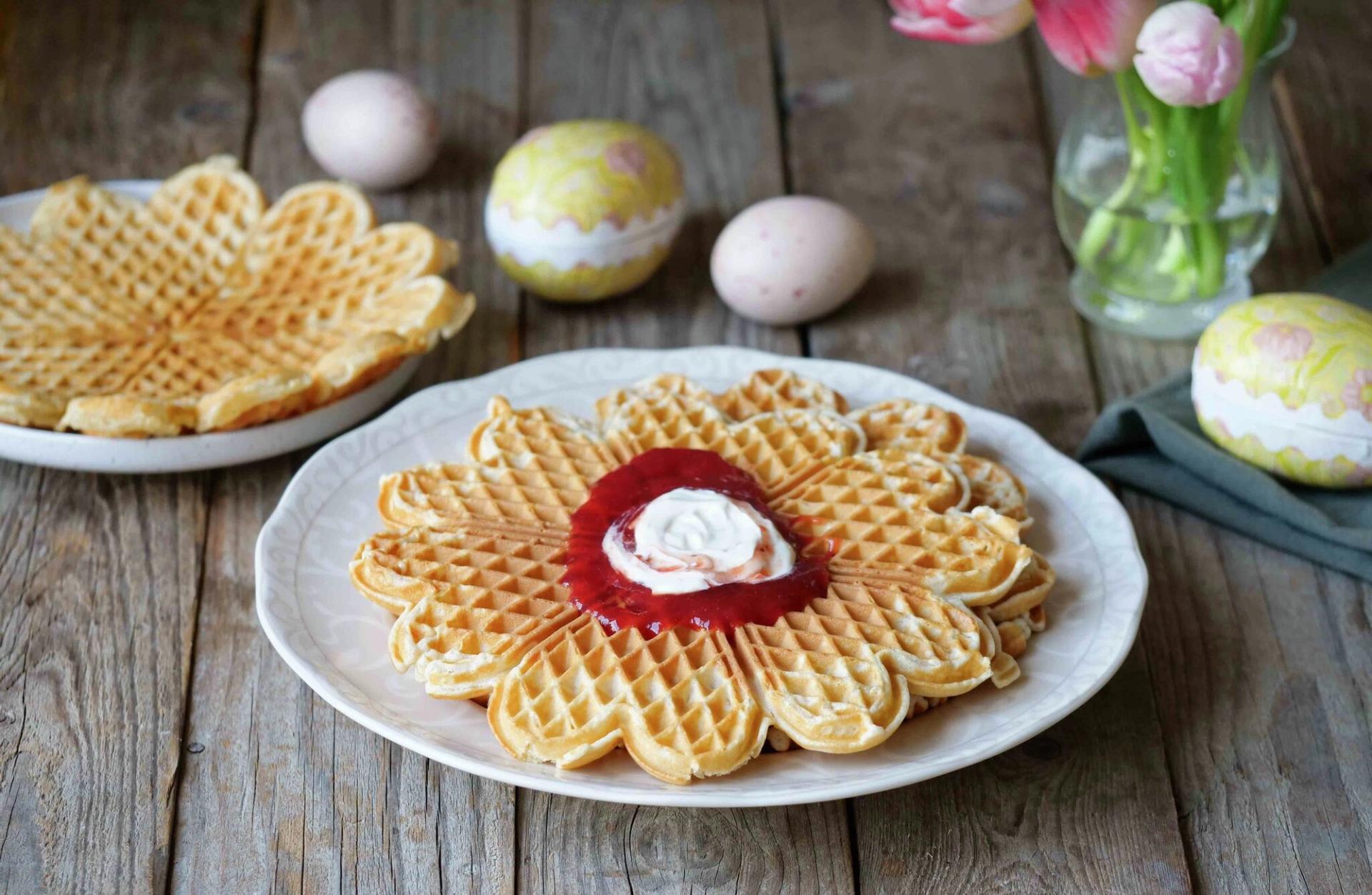 A plate of flower-shaped waffles topped with jam and cream, surrounded by Easter eggs and tulips on a wooden table.