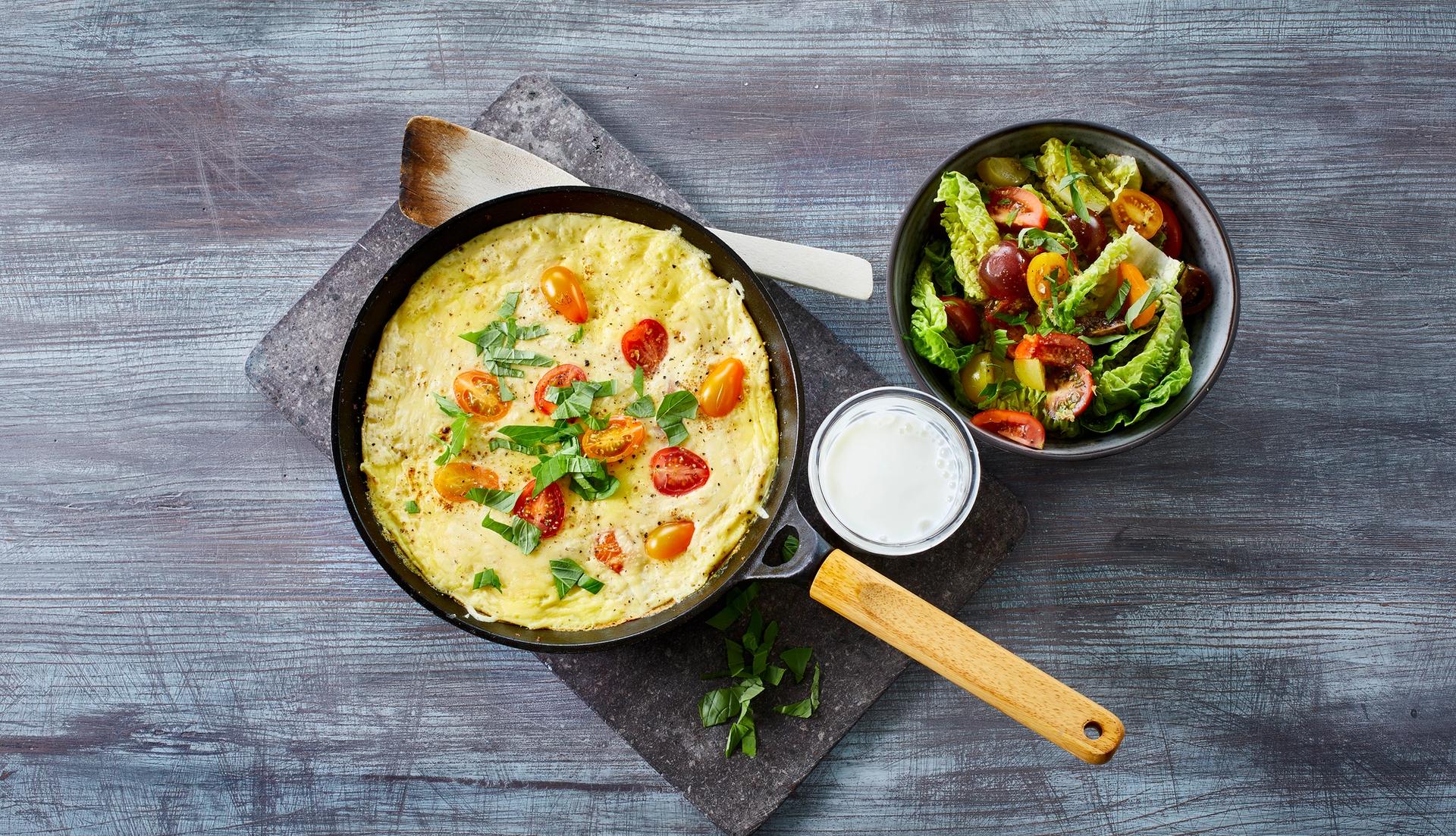 Omelett med tomater i en stekepanne med et glass melk og en skål salat ved siden