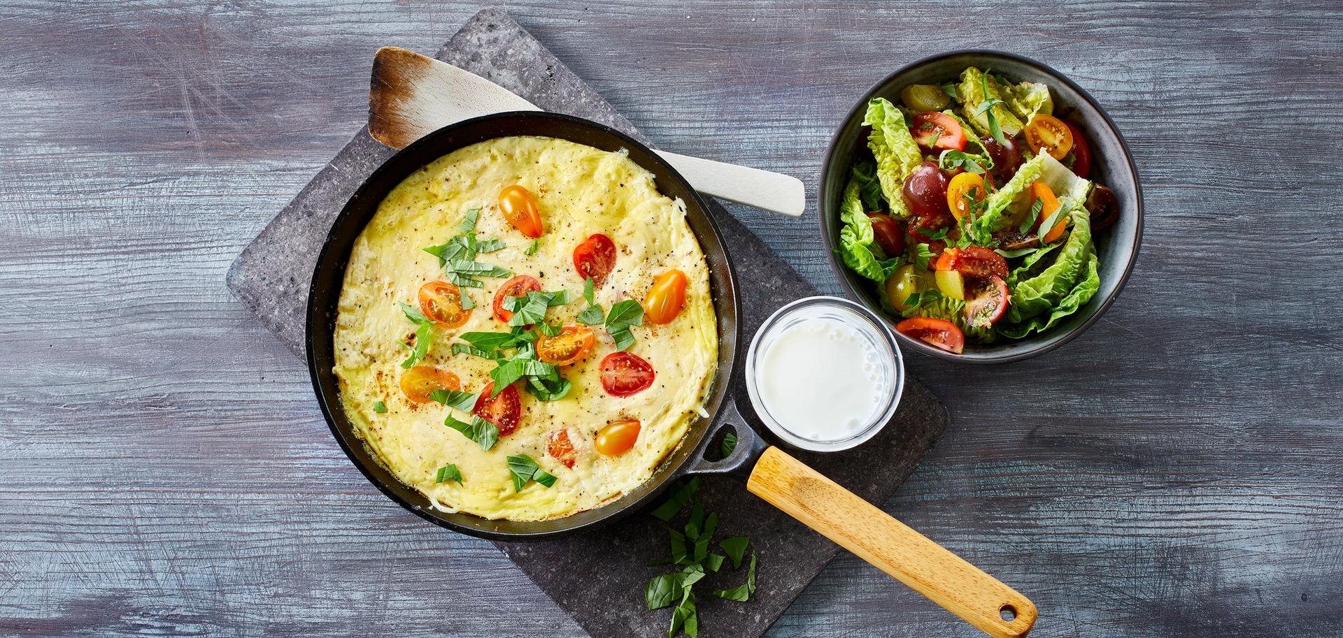 Omelett med tomater i en stekepanne med et glass melk og en skål salat ved siden