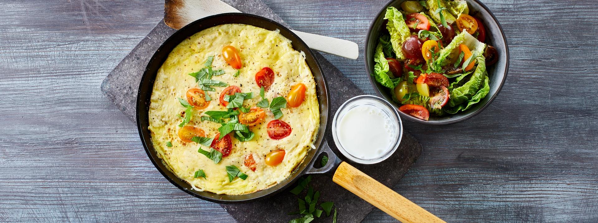 Omelett med tomater i en stekepanne med et glass melk og en skål salat ved siden