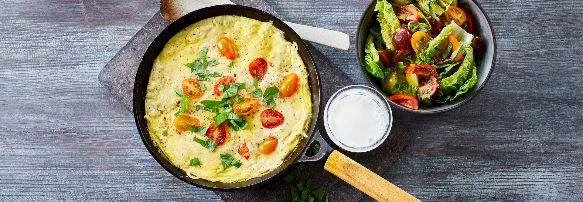 Omelett med tomater i en stekepanne med et glass melk og en skål salat ved siden