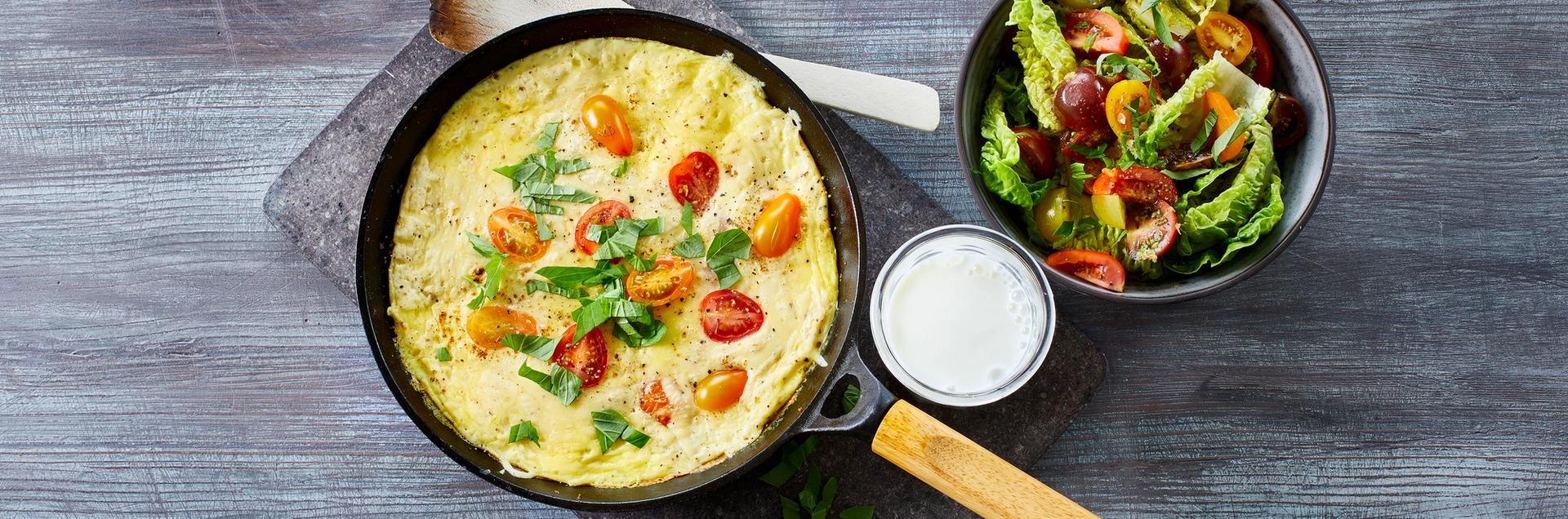 Omelett med tomater i en stekepanne med et glass melk og en skål salat ved siden