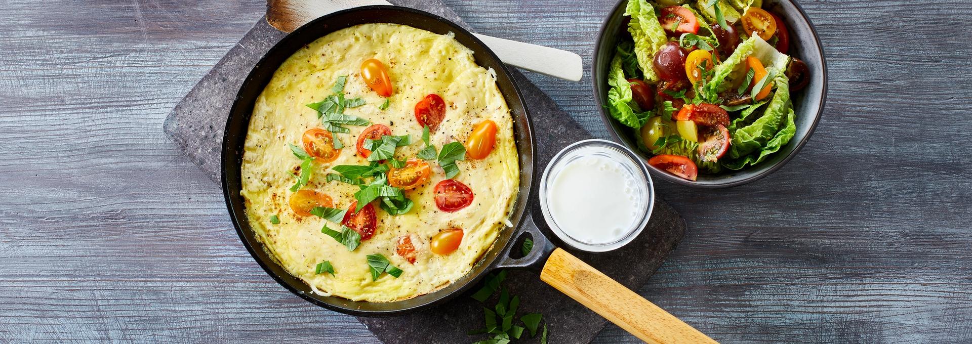 Omelett med tomater i en stekepanne med et glass melk og en skål salat ved siden