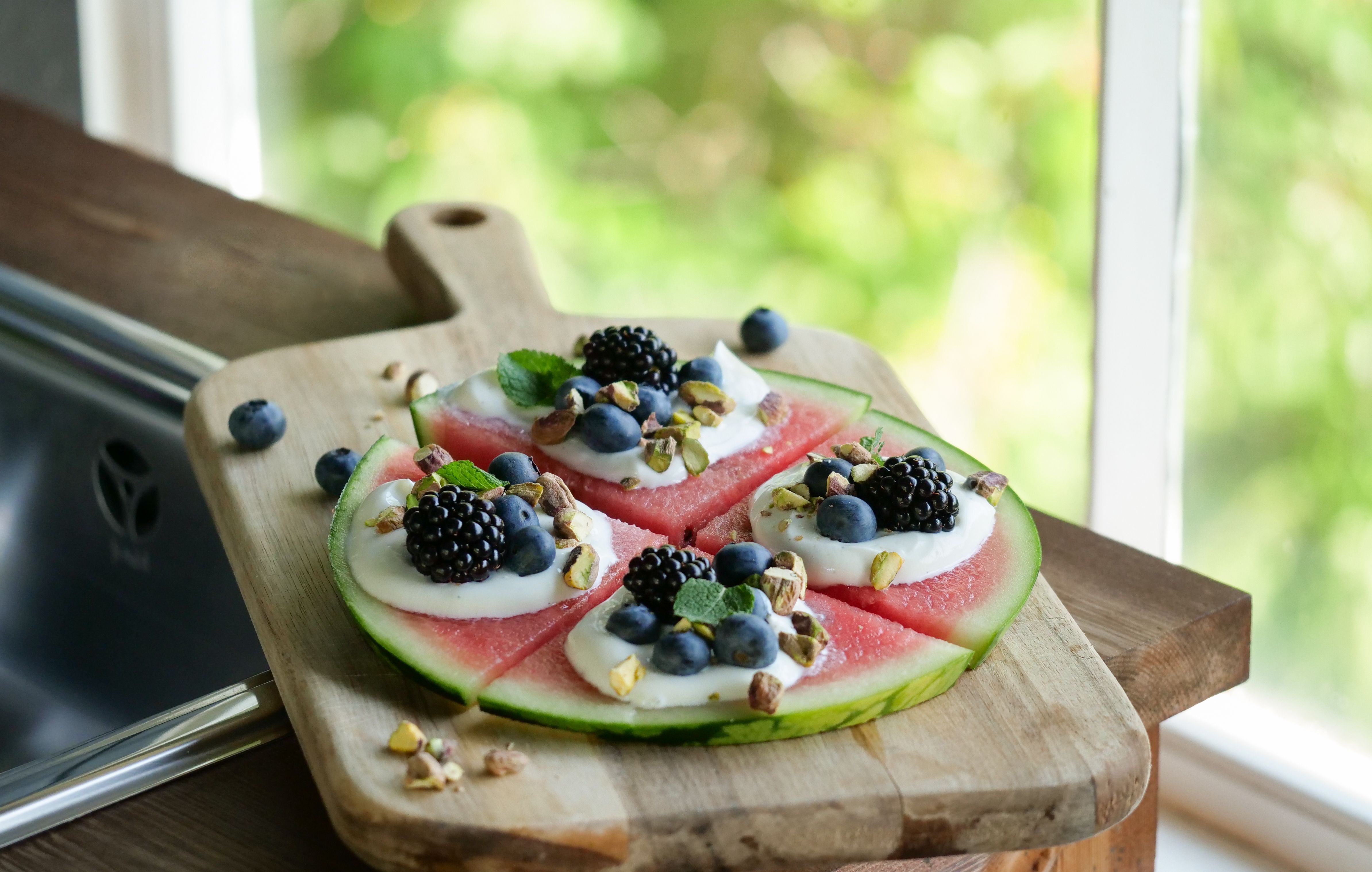 a watermelon pizza with blueberries , blackberries , pistachios and yogurt on a wooden cutting board .