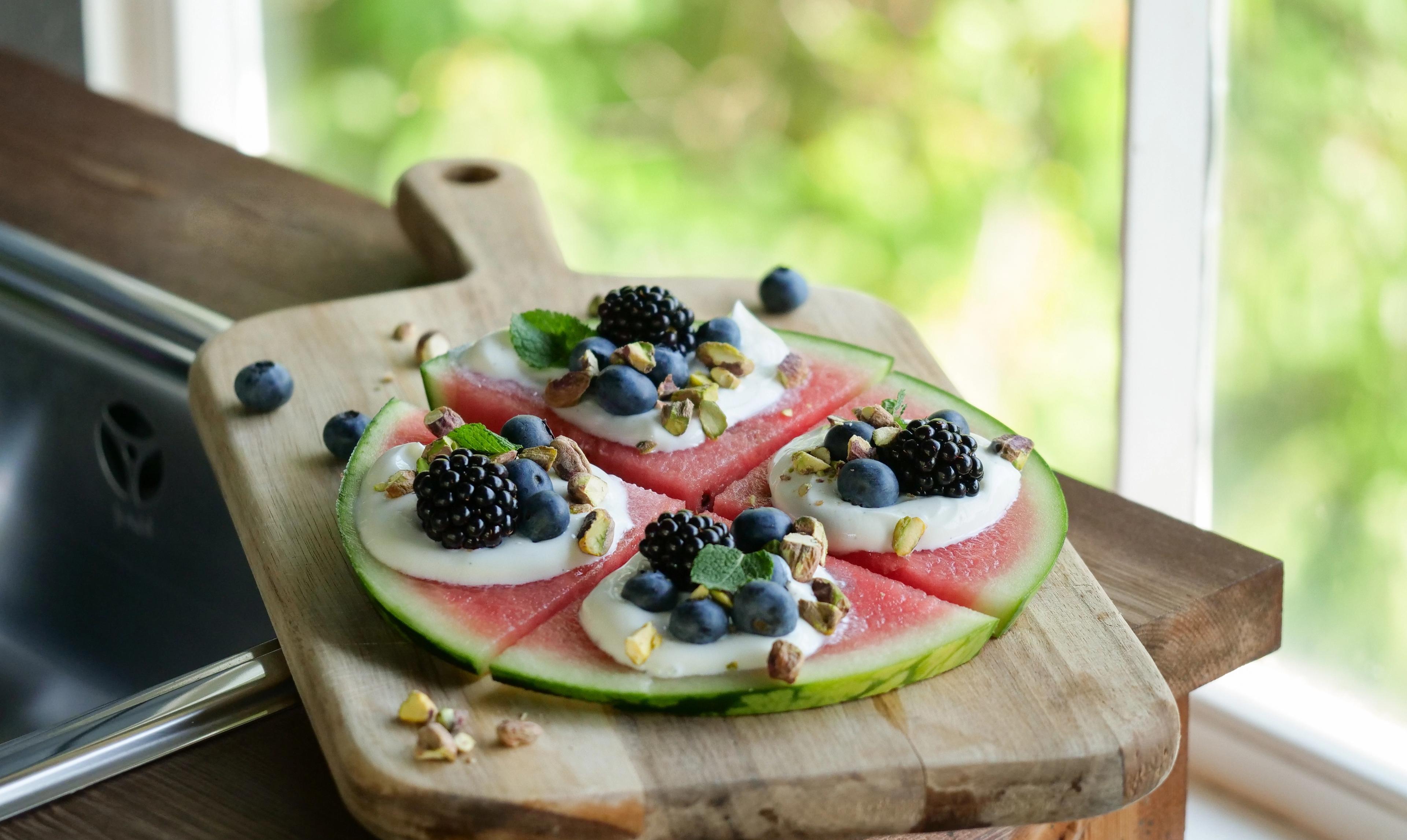 a watermelon pizza with blueberries , blackberries , pistachios and yogurt on a wooden cutting board .