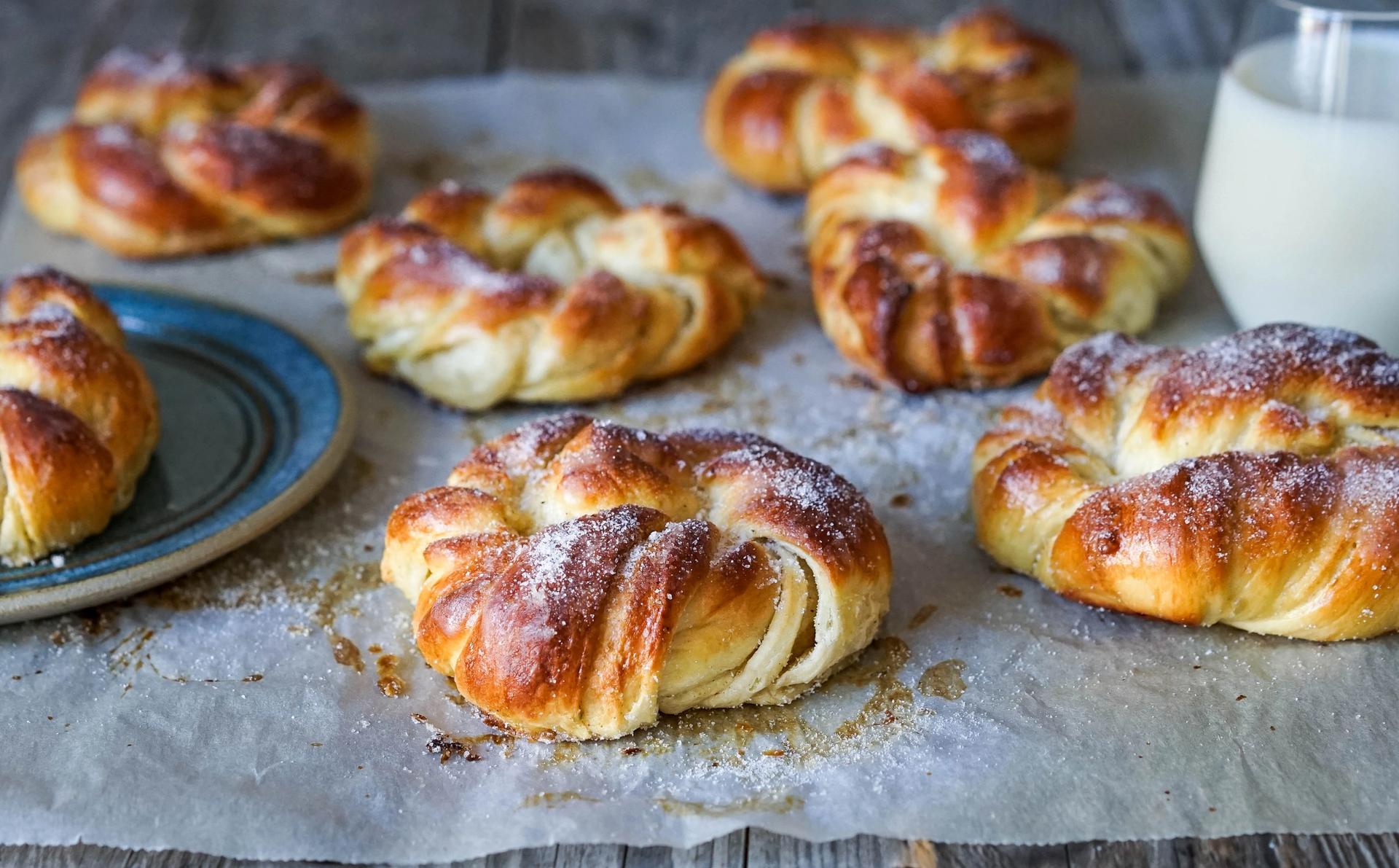 Kardemommeboller med melisdryss på et bakepapir med et glass melk ved siden av