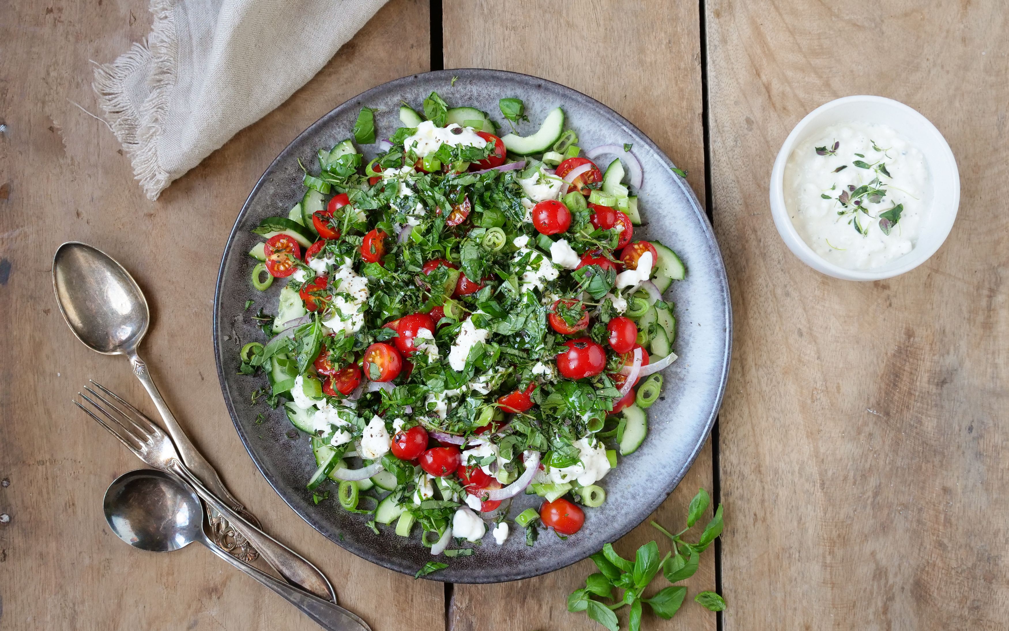Bolle med salat med tomater, agurk og cottage cheese. To skjeer, en gaffel og en skål med hvit dressing.