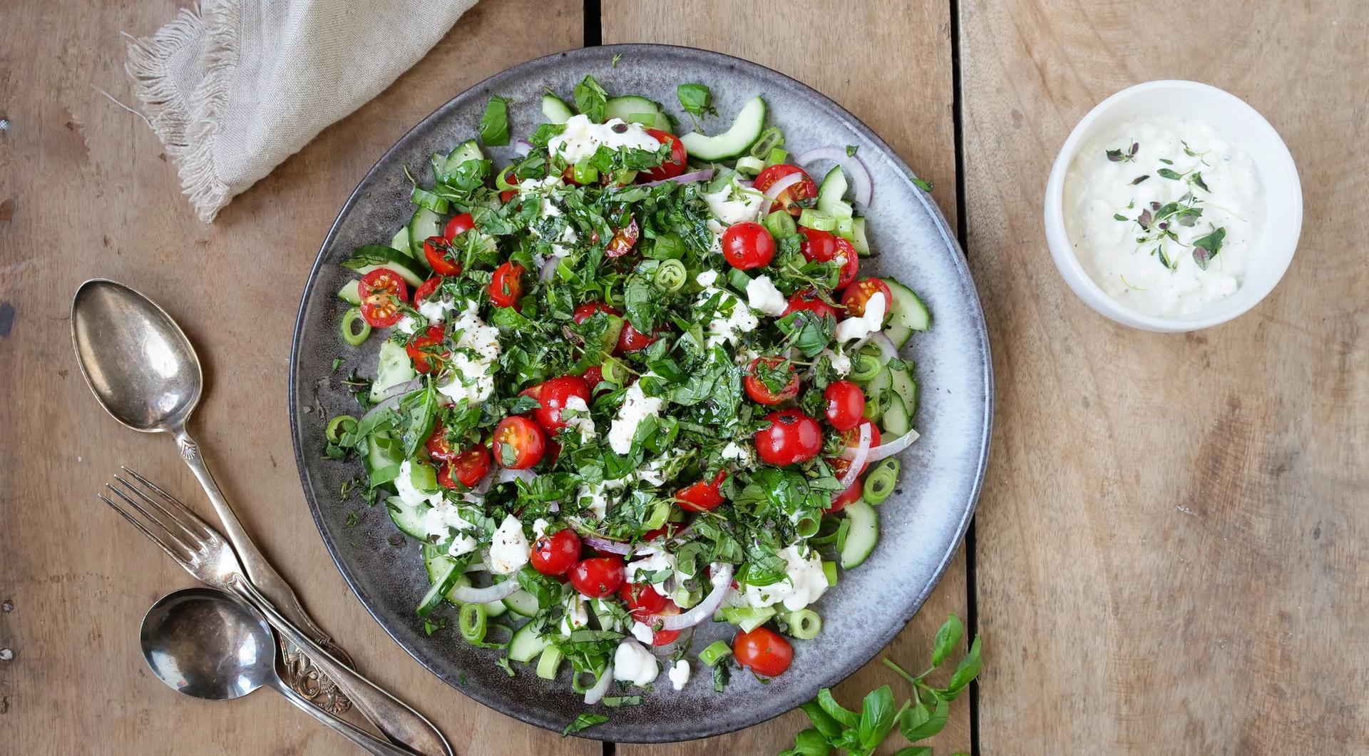 Bolle med salat med tomater, agurk og cottage cheese. To skjeer, en gaffel og en skål med hvit dressing.