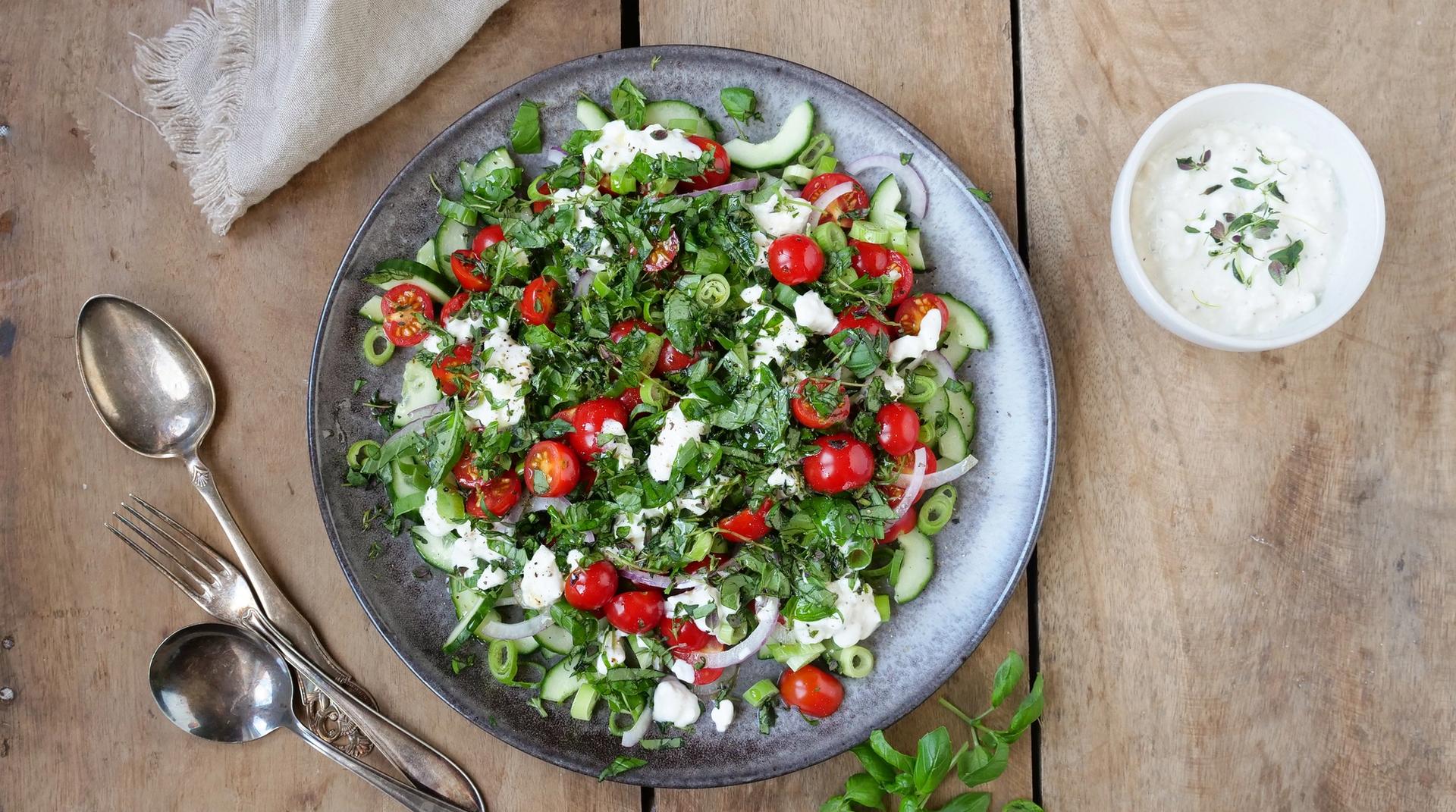 Bolle med salat med tomater, agurk og cottage cheese. To skjeer, en gaffel og en skål med hvit dressing.