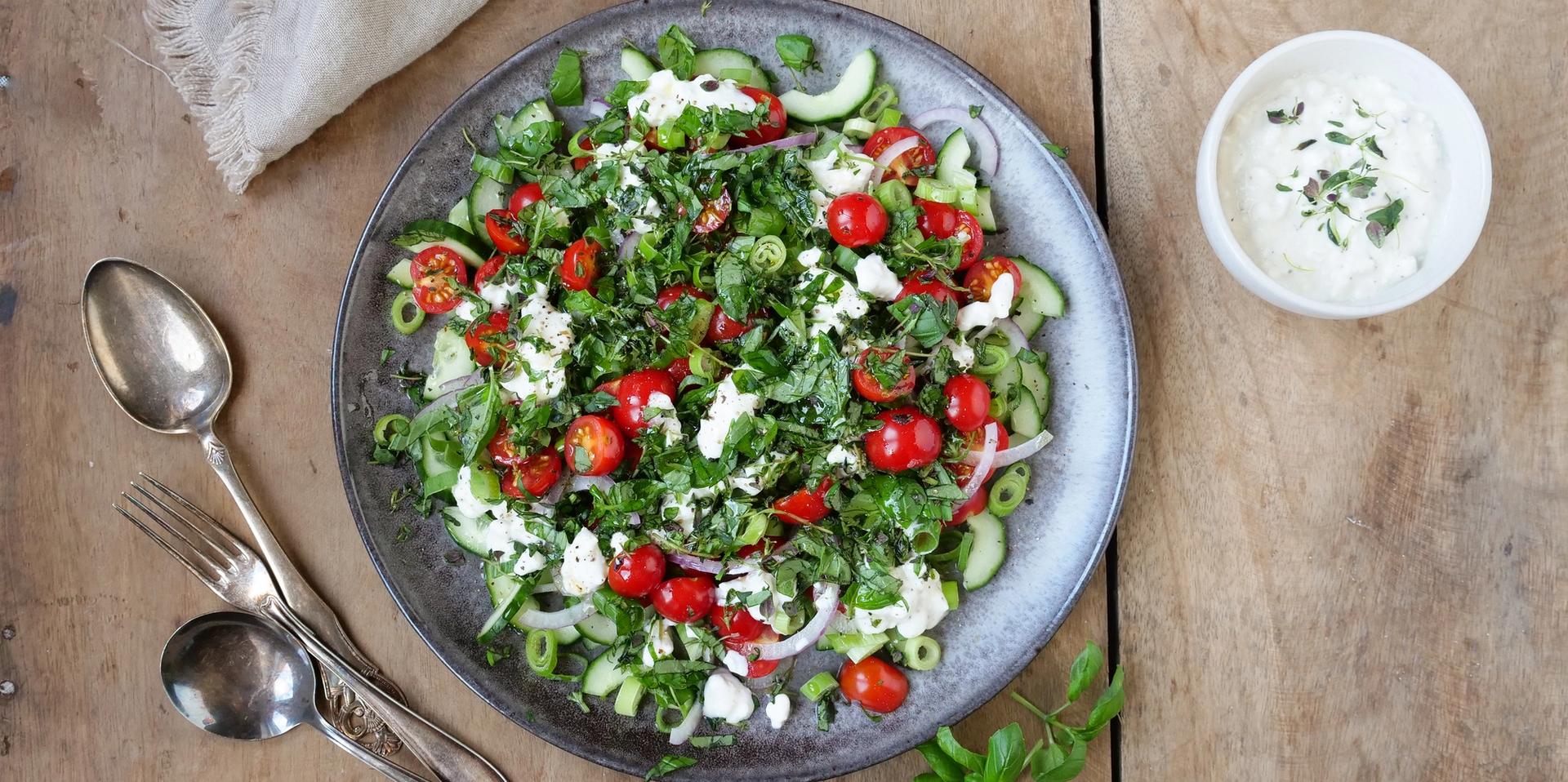 Bolle med salat med tomater, agurk og cottage cheese. To skjeer, en gaffel og en skål med hvit dressing.