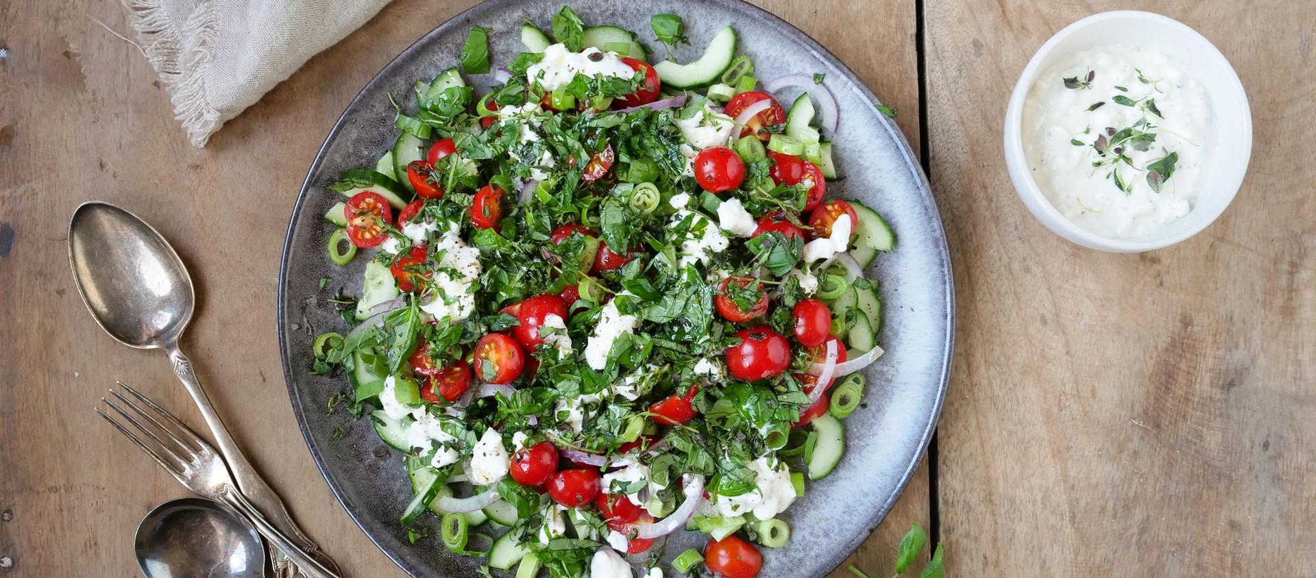 Bolle med salat med tomater, agurk og cottage cheese. To skjeer, en gaffel og en skål med hvit dressing.