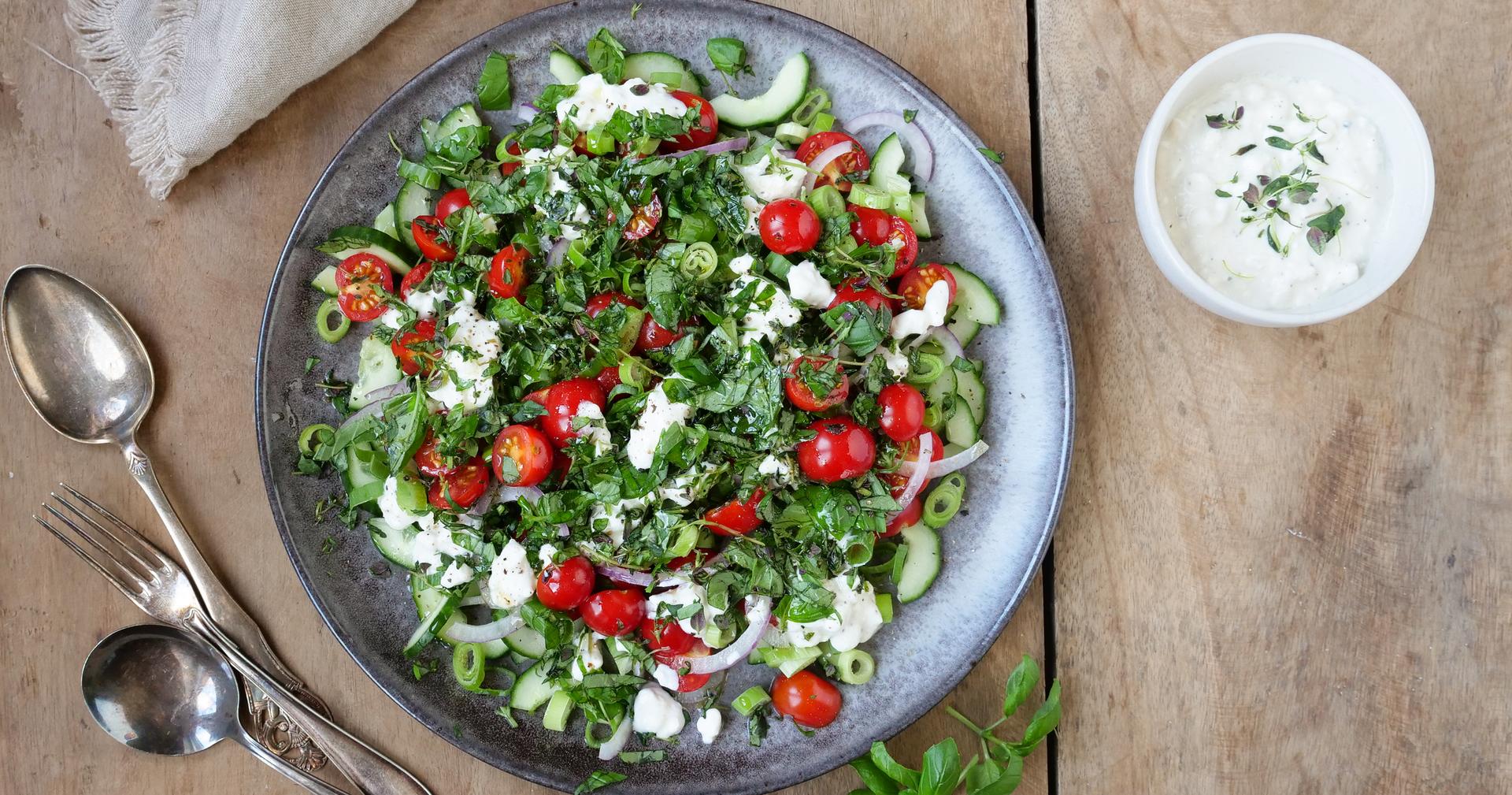 Bolle med salat med tomater, agurk og cottage cheese. To skjeer, en gaffel og en skål med hvit dressing.