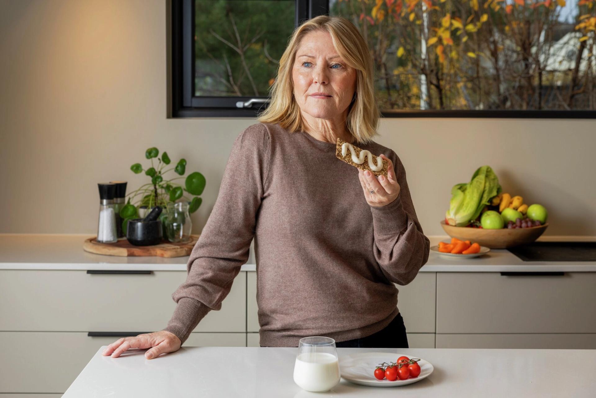 A blonde woman holds a crispbread with white topping in a modern kitchen with fruits, vegetables, and milk on the counter.