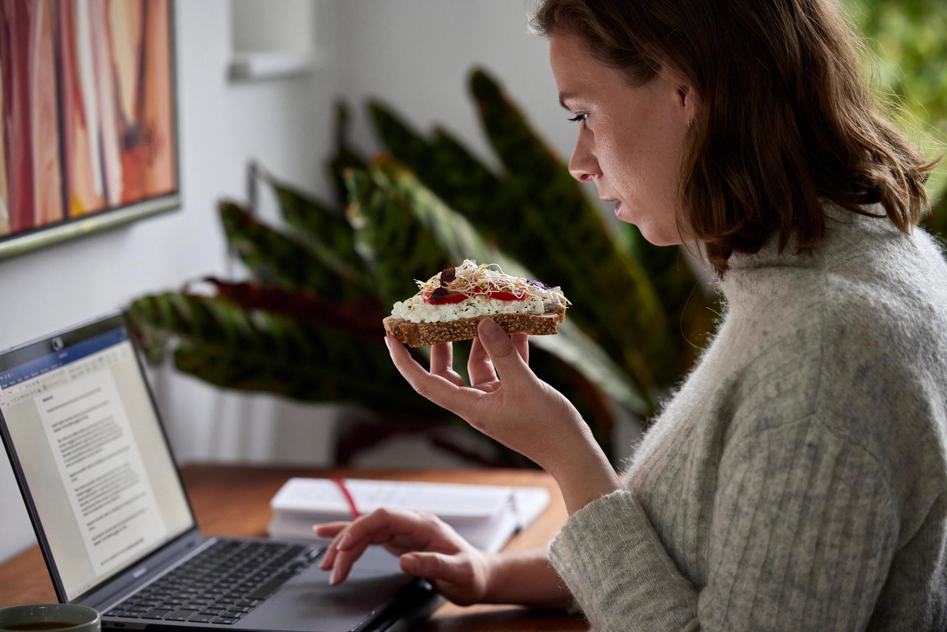 a woman is eating a sandwich while using a laptop computer .