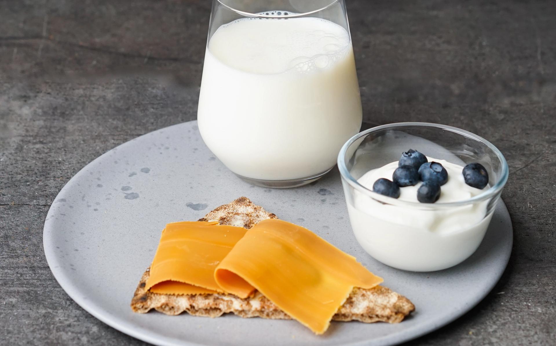 Lys grå tallerken med knekkebrød med brunost, en skål yoghurt med blåbær på toppen og et glass melk