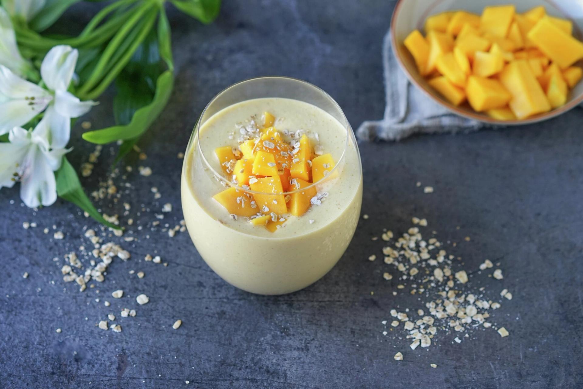 Mango smoothie in a glass, topped with diced mango and oats, with a bowl of mango chunks and white flowers in the background.