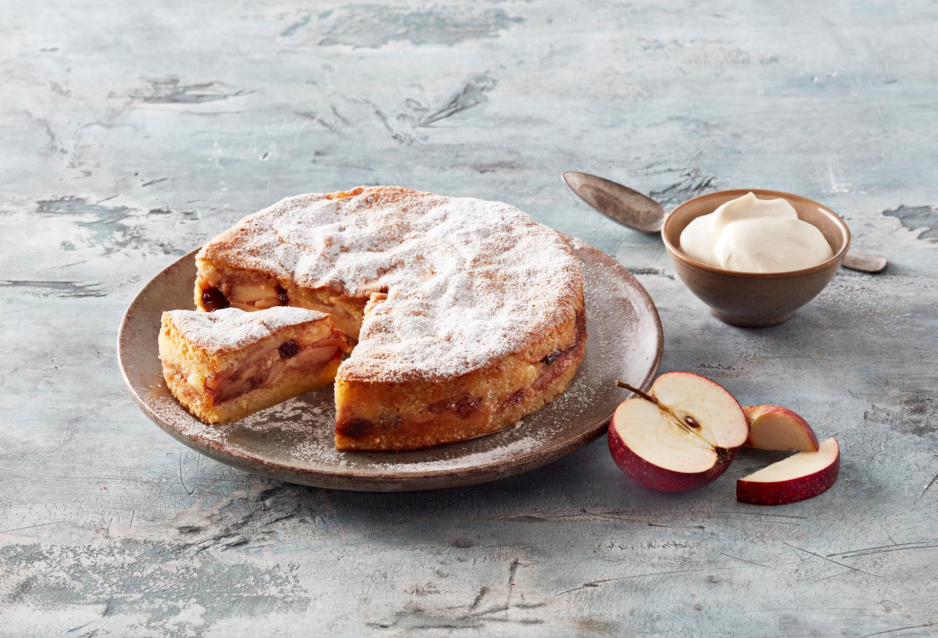 Apple cake with a slice removed, dusted with powdered sugar, served with sliced apples and cream.
