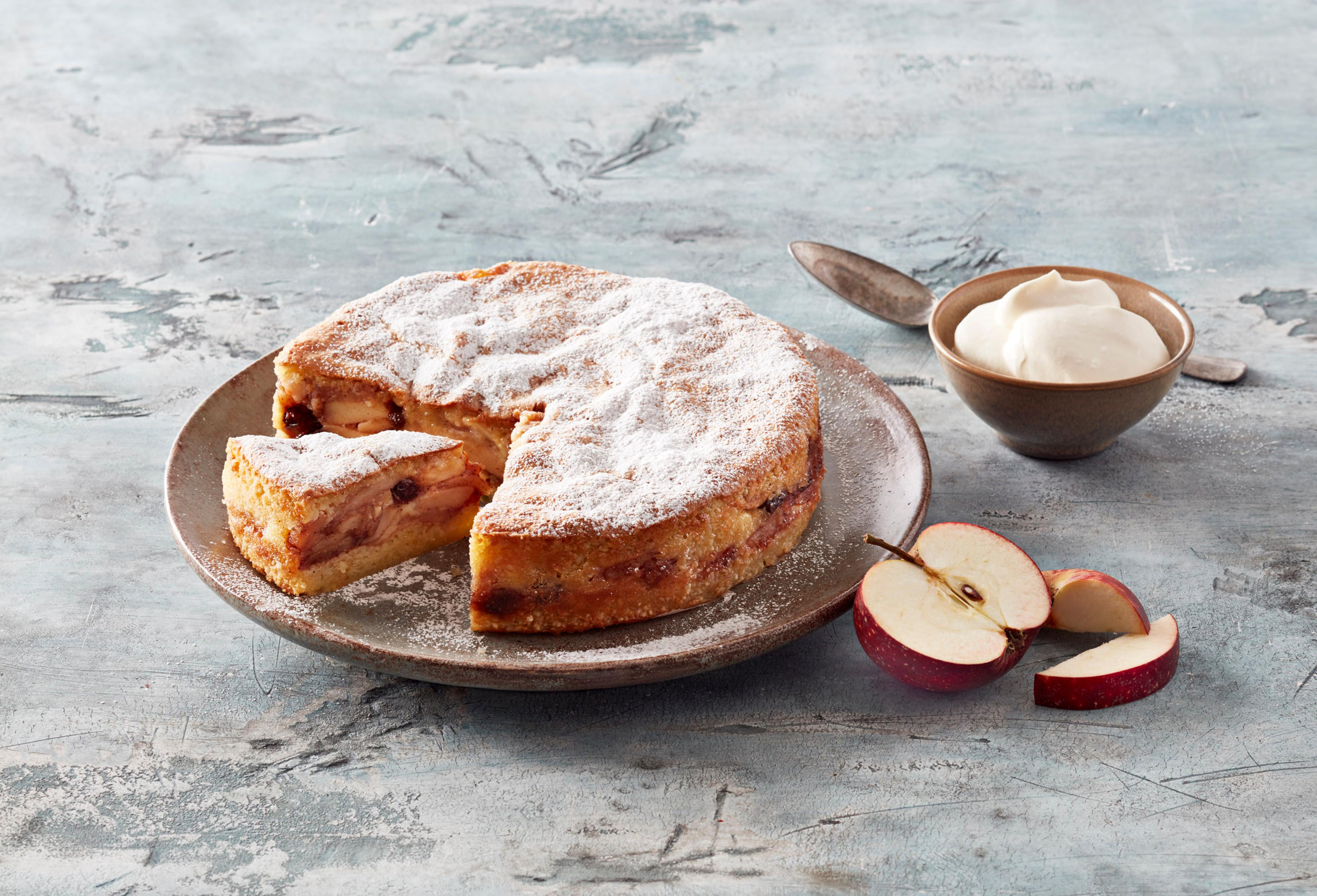 Apple cake with a slice removed, dusted with powdered sugar, served with sliced apples and cream.