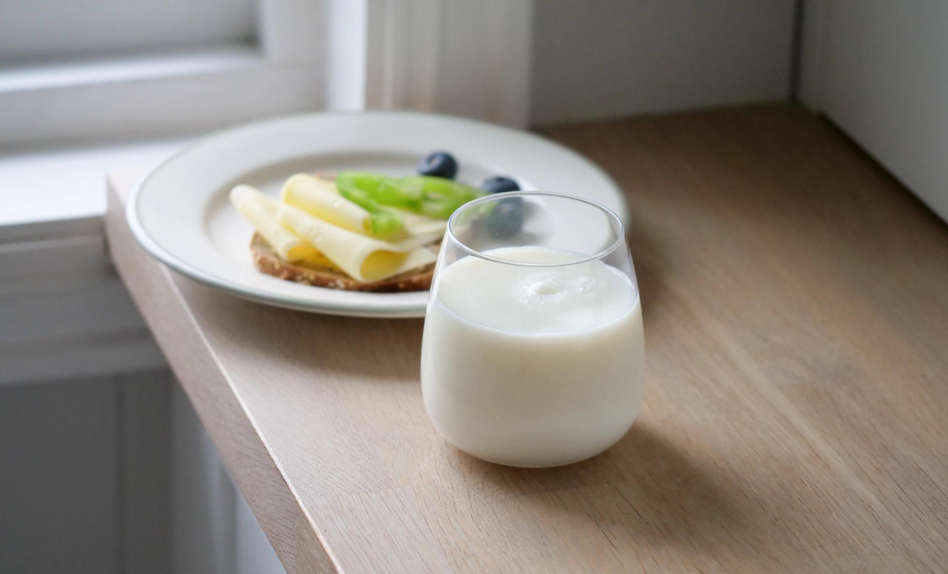 A glass of milk and a plate with cheese, kiwi, and blueberries on a cracker, on a wooden windowsill.