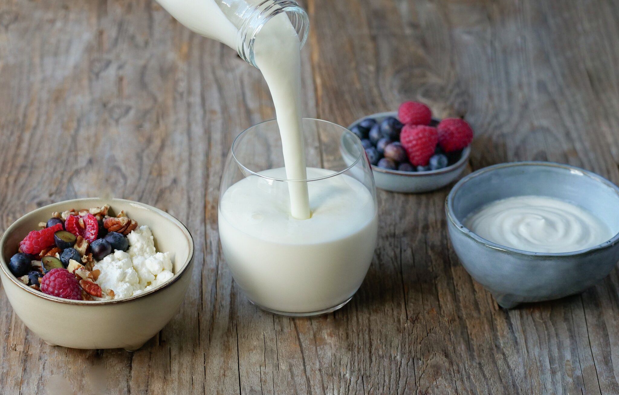 Milk poured into a glass, with cottage cheese, berries, nuts, and yogurt on a wooden table.