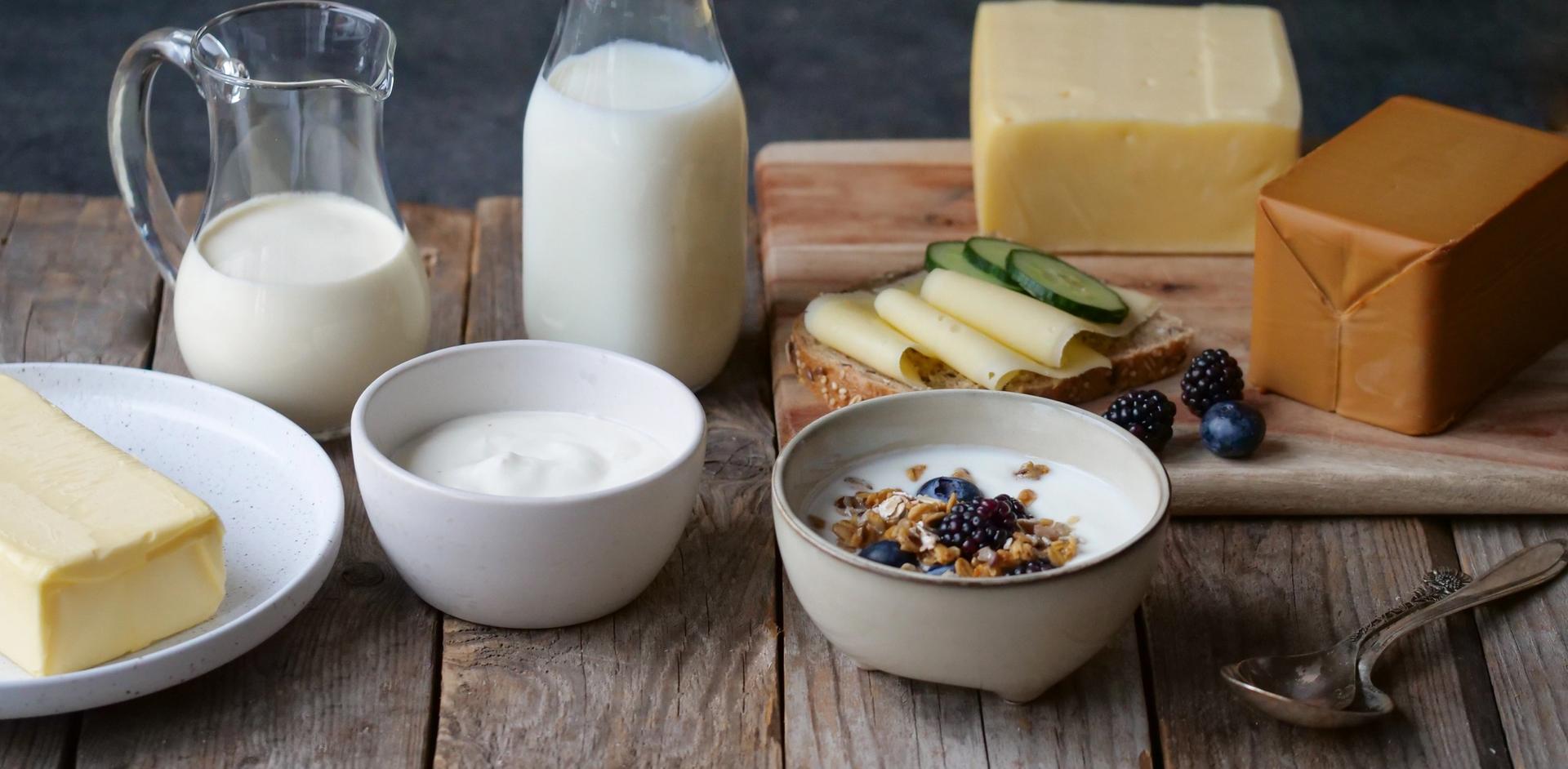 En rustikk fremstilling av melk, smør, gulost, brunost, yoghurt, frokostblanding med melk og fløte i en glasskanne.