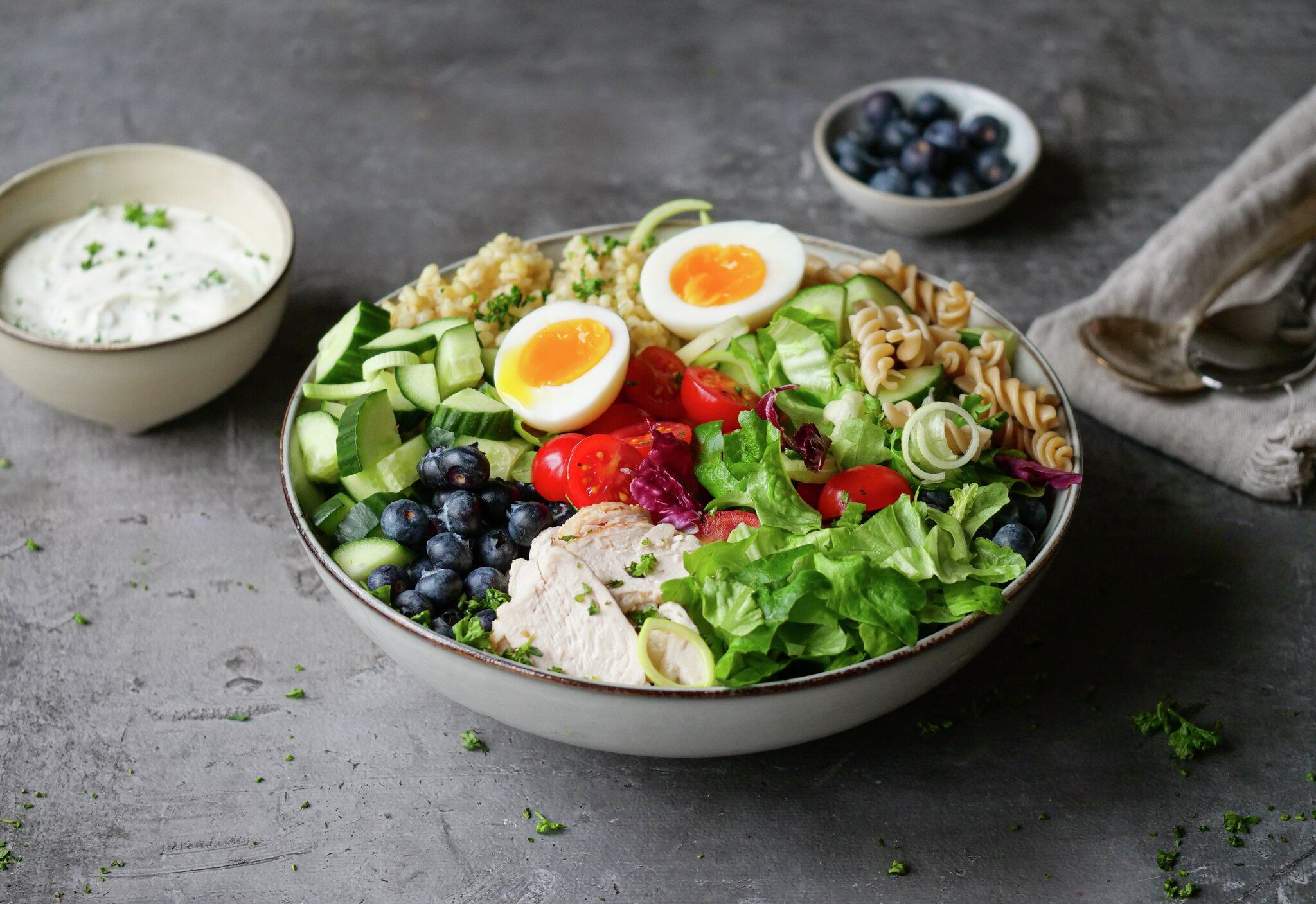 A vibrant salad bowl with sliced chicken, halved soft-boiled eggs, blueberries, mixed greens, fresh vegetables, and grains, served with a side of creamy white dressing.