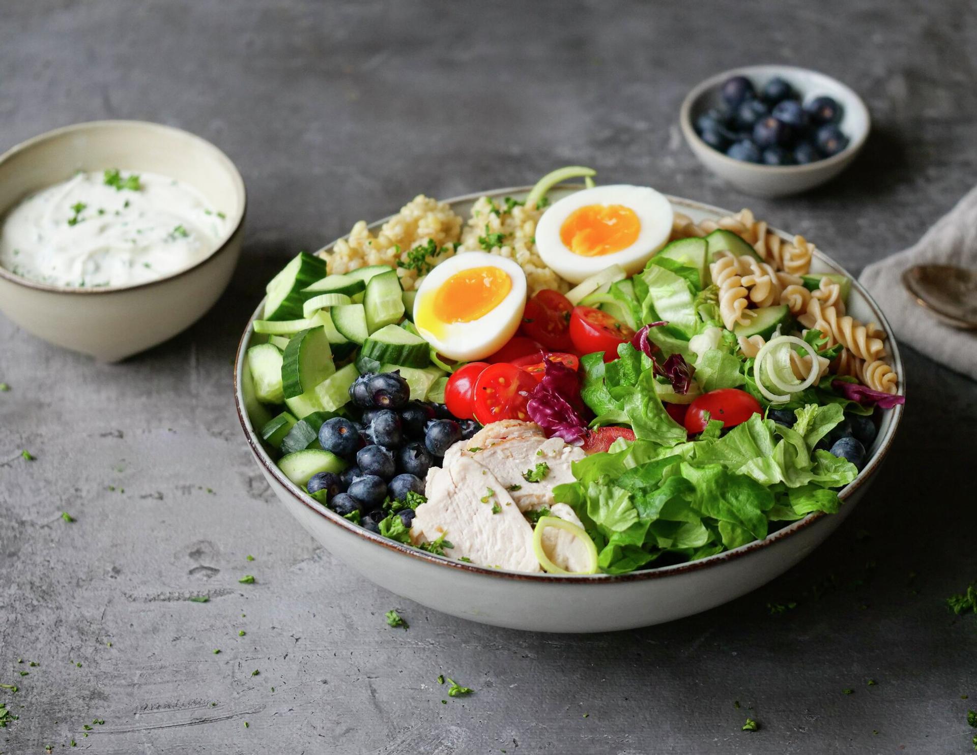 A colorful bowl of salad with chicken, soft-boiled eggs, blueberries, mixed vegetables, and pasta, served with a side of creamy dressing.
