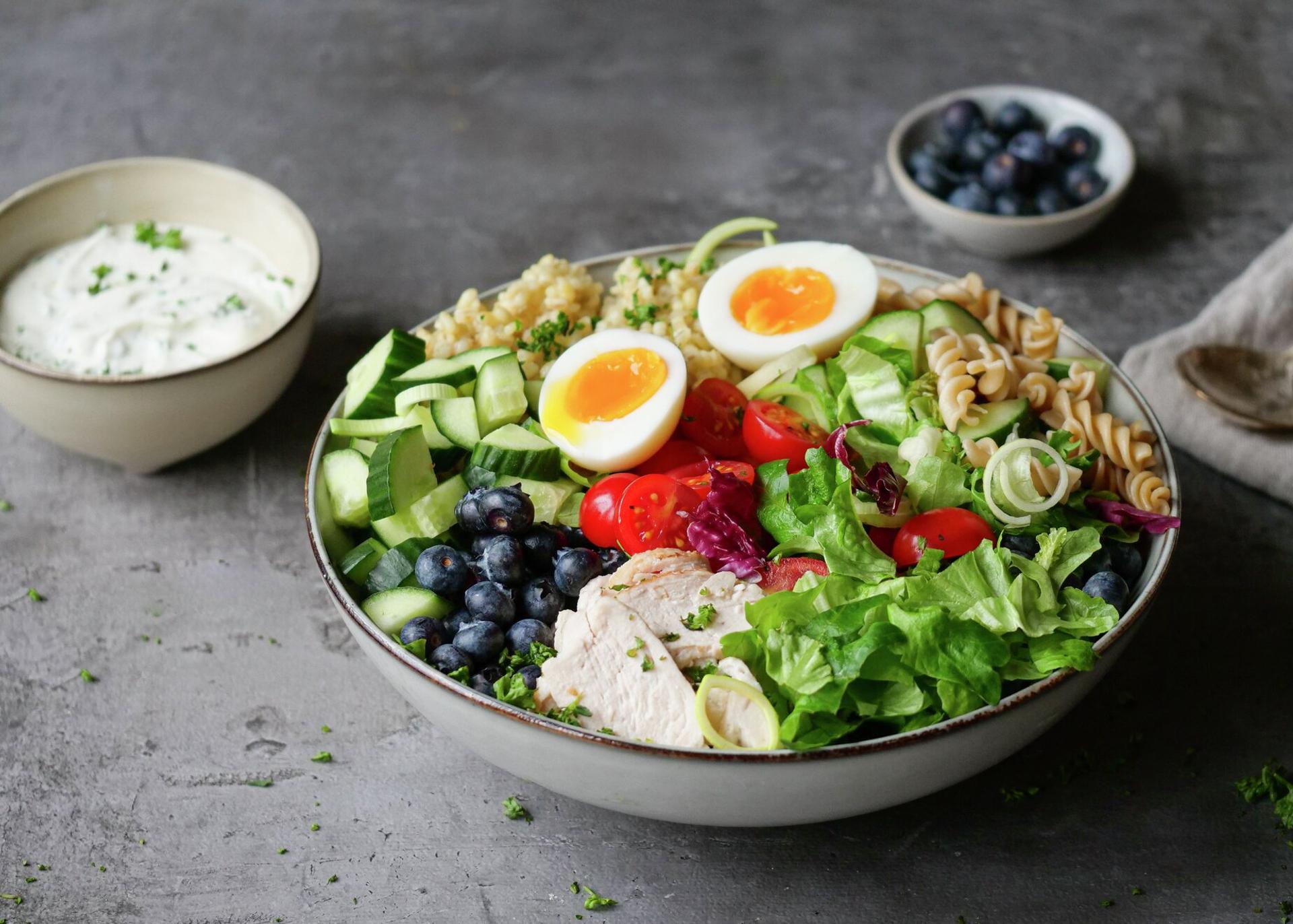 A vibrant salad bowl with sliced chicken, halved soft-boiled eggs, blueberries, mixed greens, fresh vegetables, and grains, served with a side of creamy white dressing.