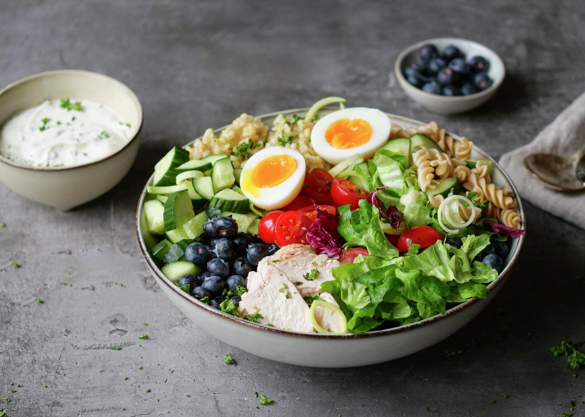 A vibrant salad bowl with chicken, soft-boiled eggs, blueberries, cucumber, tomatoes, pasta, and mixed greens, with a side of creamy dressing.
