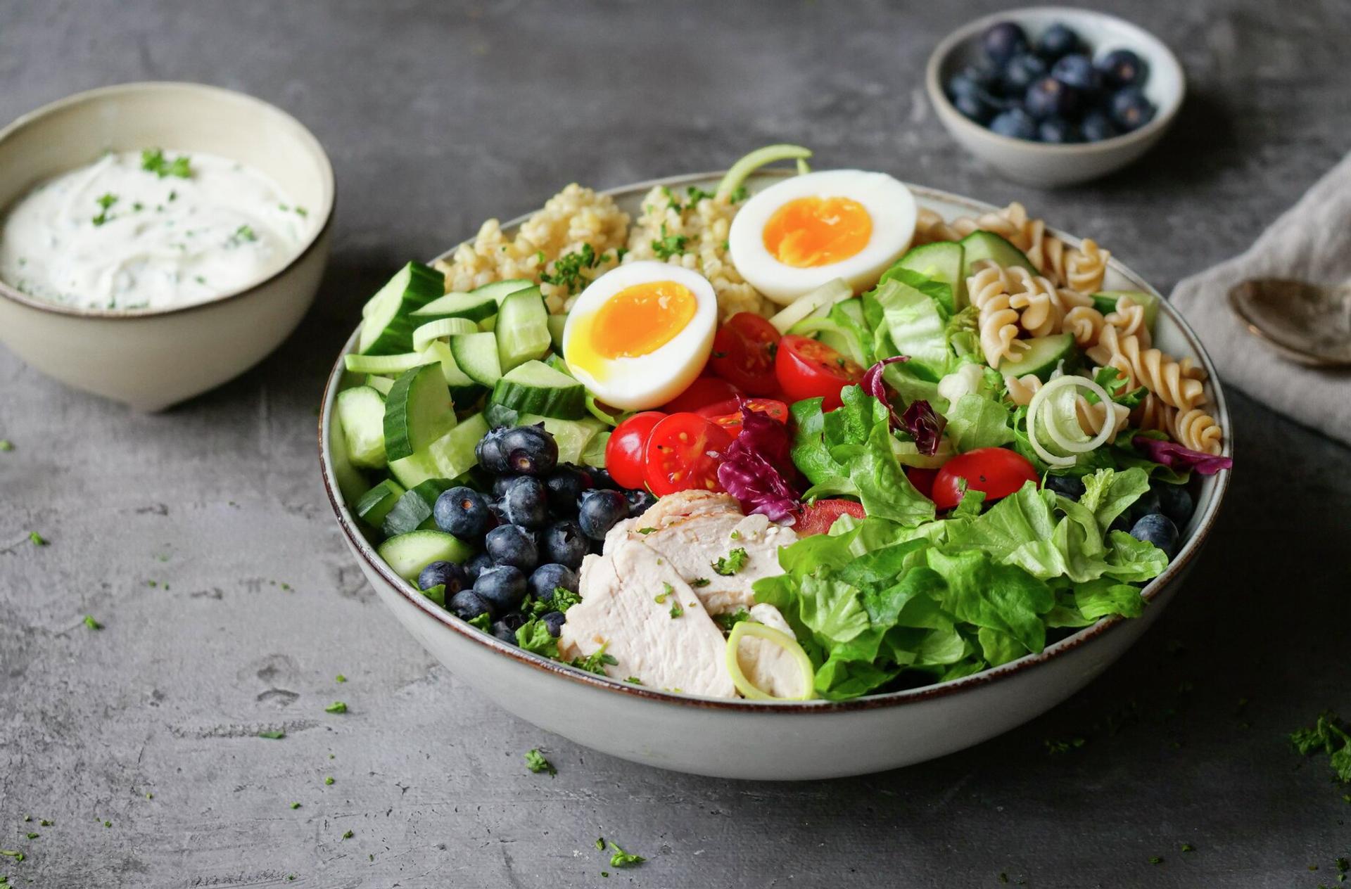 A healthy meal bowl with soft-boiled eggs, chicken, blueberries, mixed vegetables, pasta, and grains, served with a side dressing.