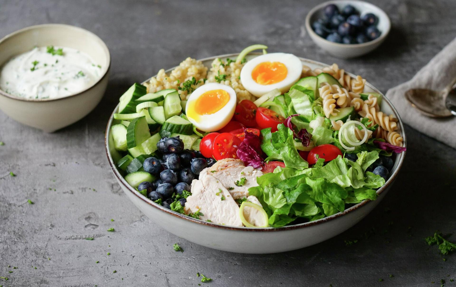 A colorful bowl with sliced chicken, two soft-boiled eggs, blueberries, cucumber, tomatoes, mixed greens, pasta, and quinoa, served with a side of creamy dressing.