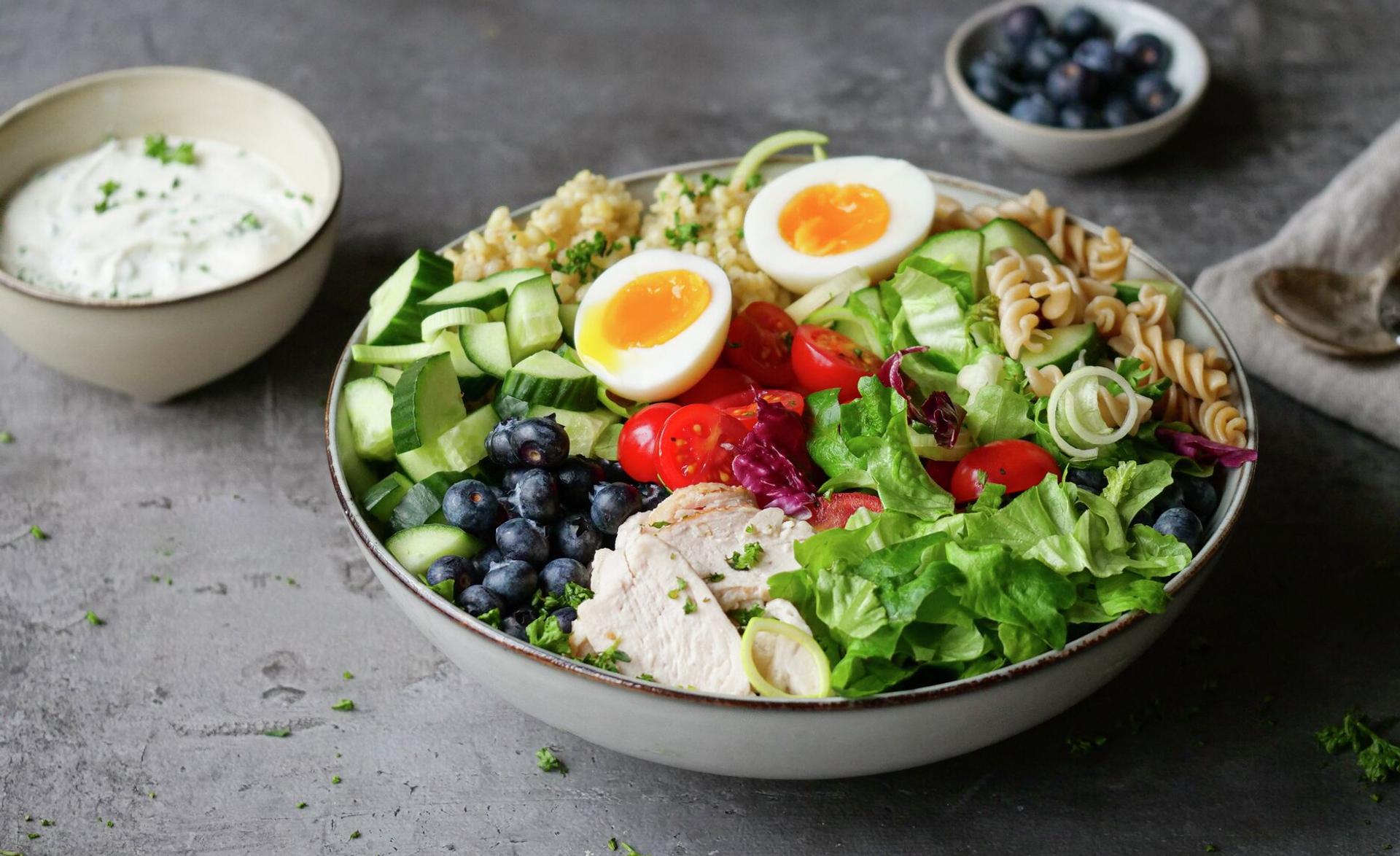 A colorful meal bowl with soft-boiled eggs, chicken, fresh vegetables, blueberries, grain, and pasta, with a side of dressing.