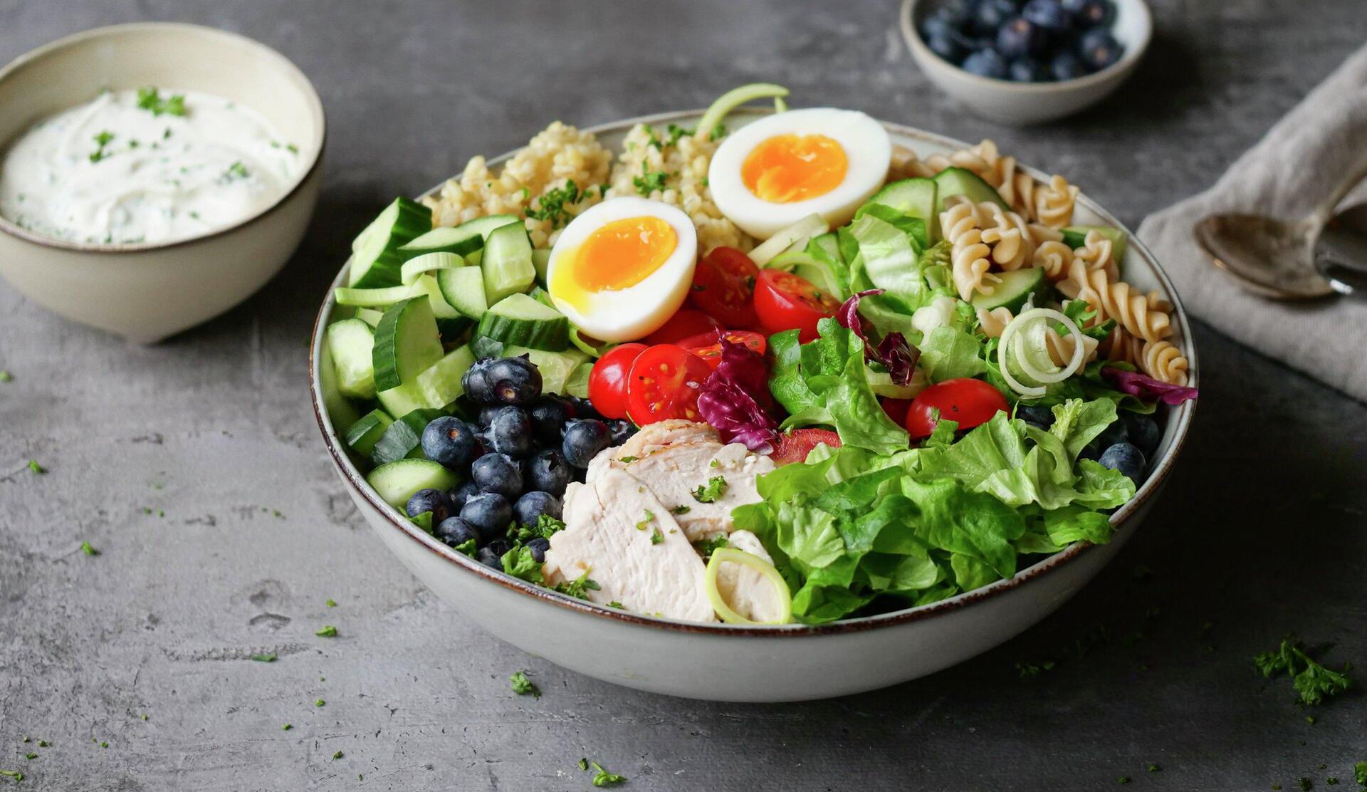 A large bowl filled with sliced chicken, soft-boiled eggs, blueberries, cucumber, cherry tomatoes, mixed greens, quinoa, and fusilli pasta, with a side of white dressing.